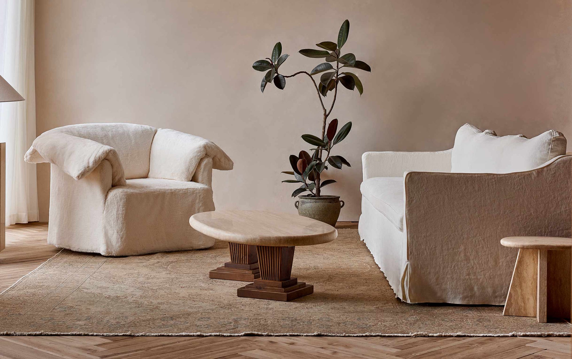 Cordelia Coffee Table in Turkish Galata Travertine in a warm beige with gold and pink undertones. Table legs made from 100% Reclaimed Chinese Pine. Table is places in a naturally lit room between a Loula Chair and an Esmé Sofa on a muted golden rug, with a tall potted plant in the background.