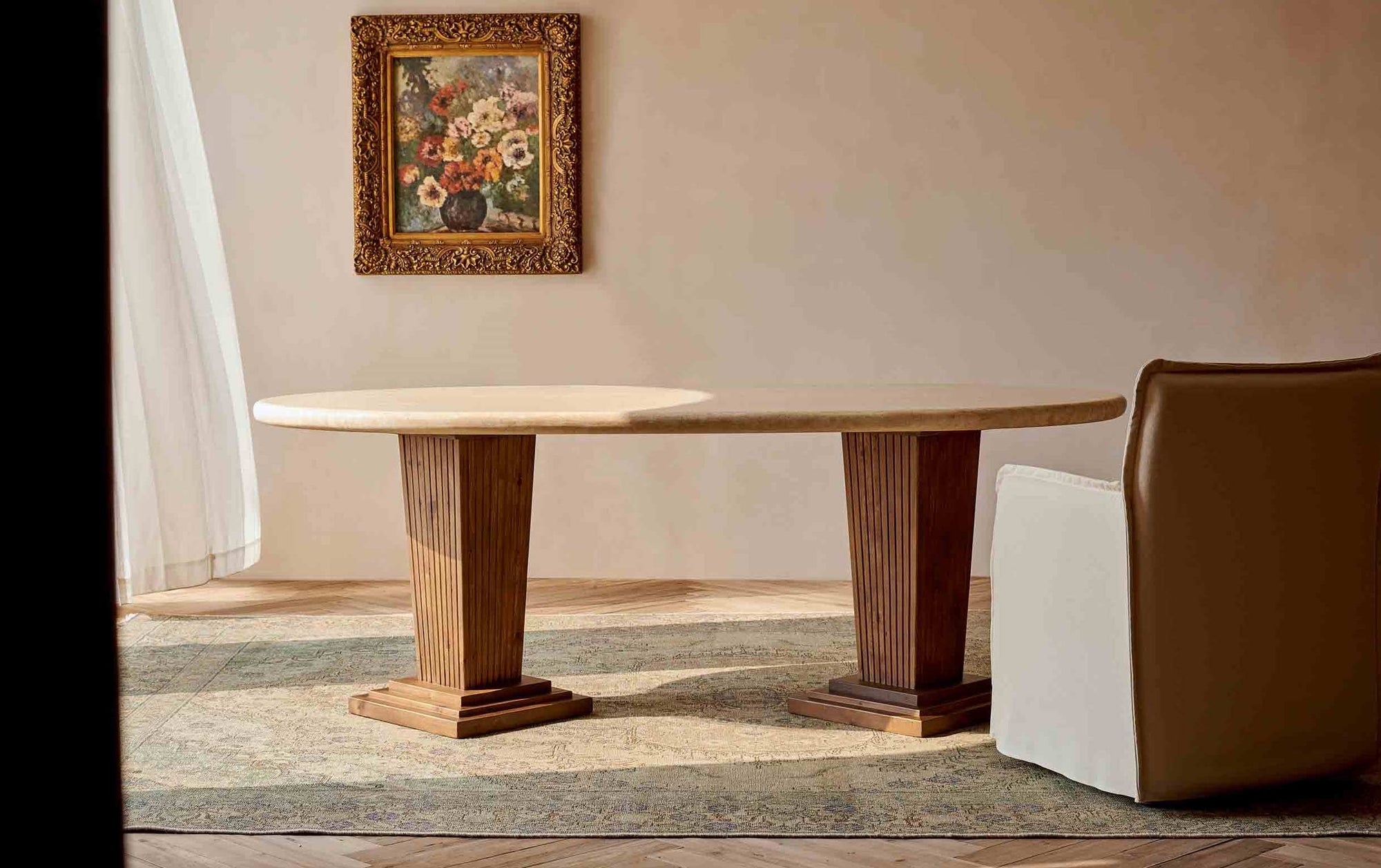 Cordelia Dining Table placed in a sunny room with a Melo Dining Chair and a framed painting. Tabletop made of Turkish Galata Travertine in a warm beige with gold and pink undertones. Various veining, color variations, and pitting throughout. Legs made of 100% Reclaimed Chinese Pine