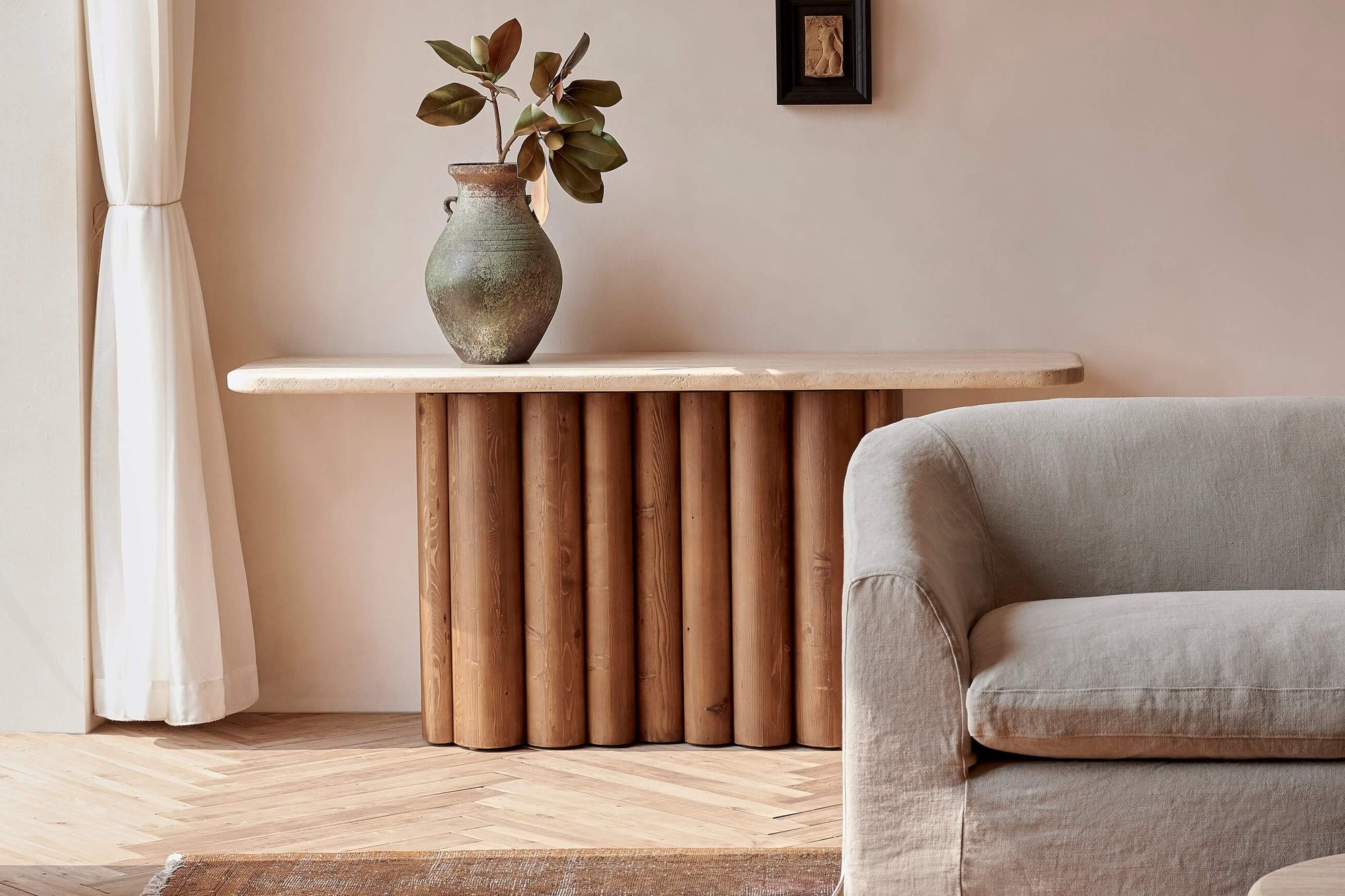 Enzo Console Table in Turkish Galata Travertine in a warm beige with gold and pink undertones. Various veining, color variations, and pitting throughout. Table base made from 100% Reclaimed Chinese Pine. Console is placed against a wall behind a Ziki Sofa, and decorated with a plant in a green vase.