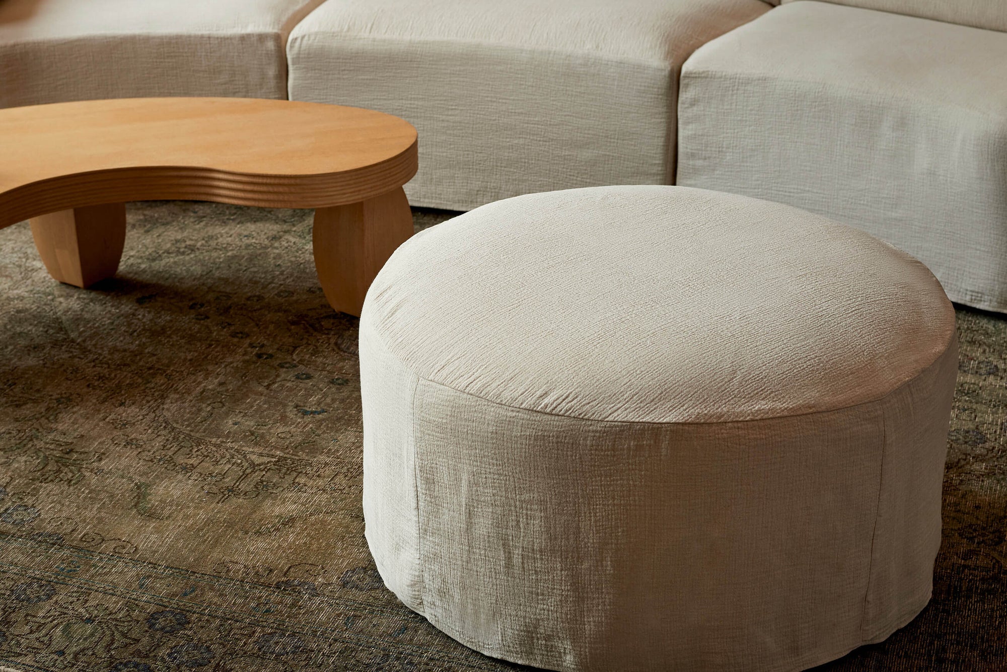 Loula Ottoman in Corn Silk, a light beige Washed Cotton Linen, placed in the foreground of a room with the Loula Sectional Sofa and Pisces Coffee Table in the background