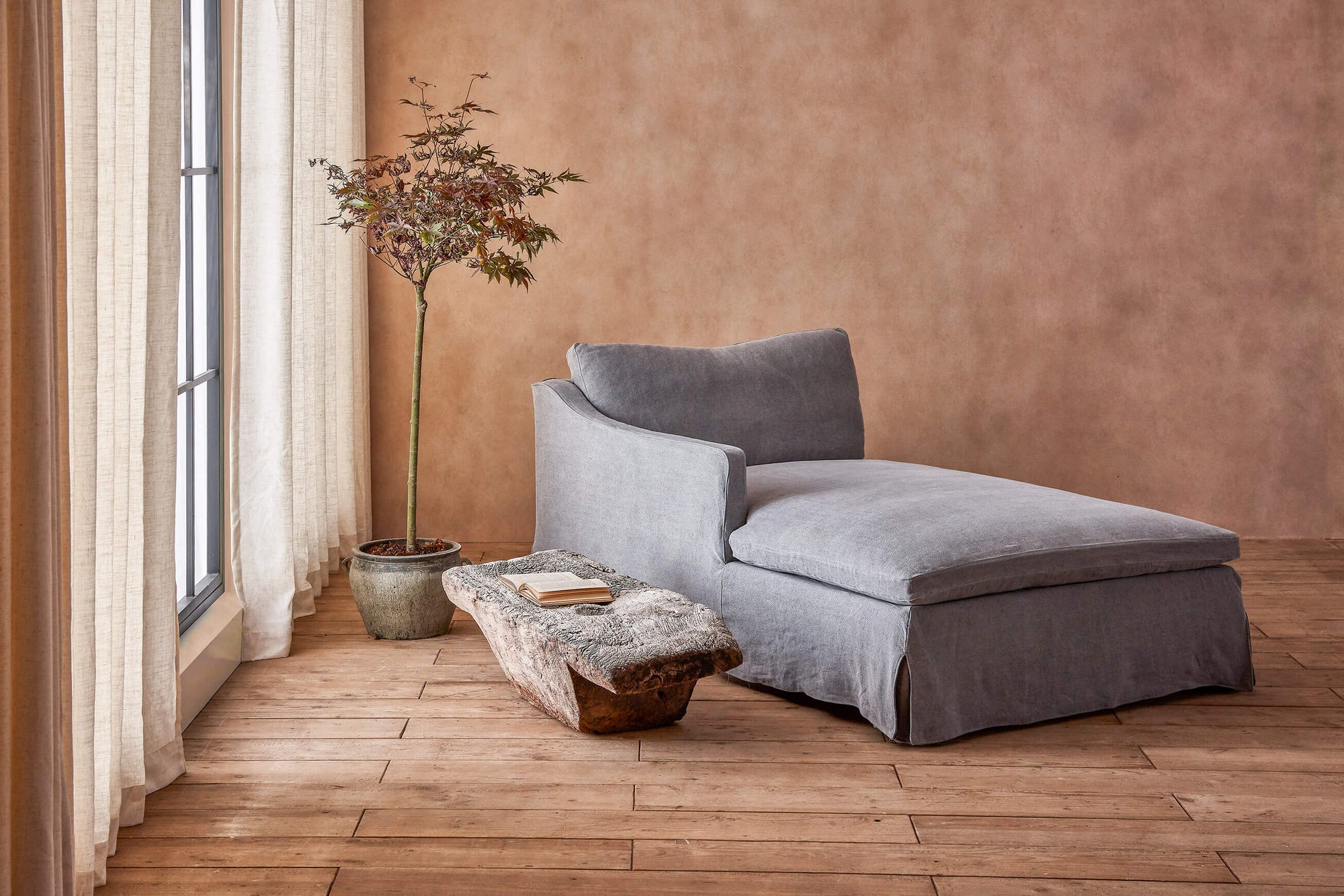 Amelia Left Arm Facing Daybed in Ink Cap, a medium cool grey Light Weight Linen, placed in a warmly lit room next to a stone table and a potted tree