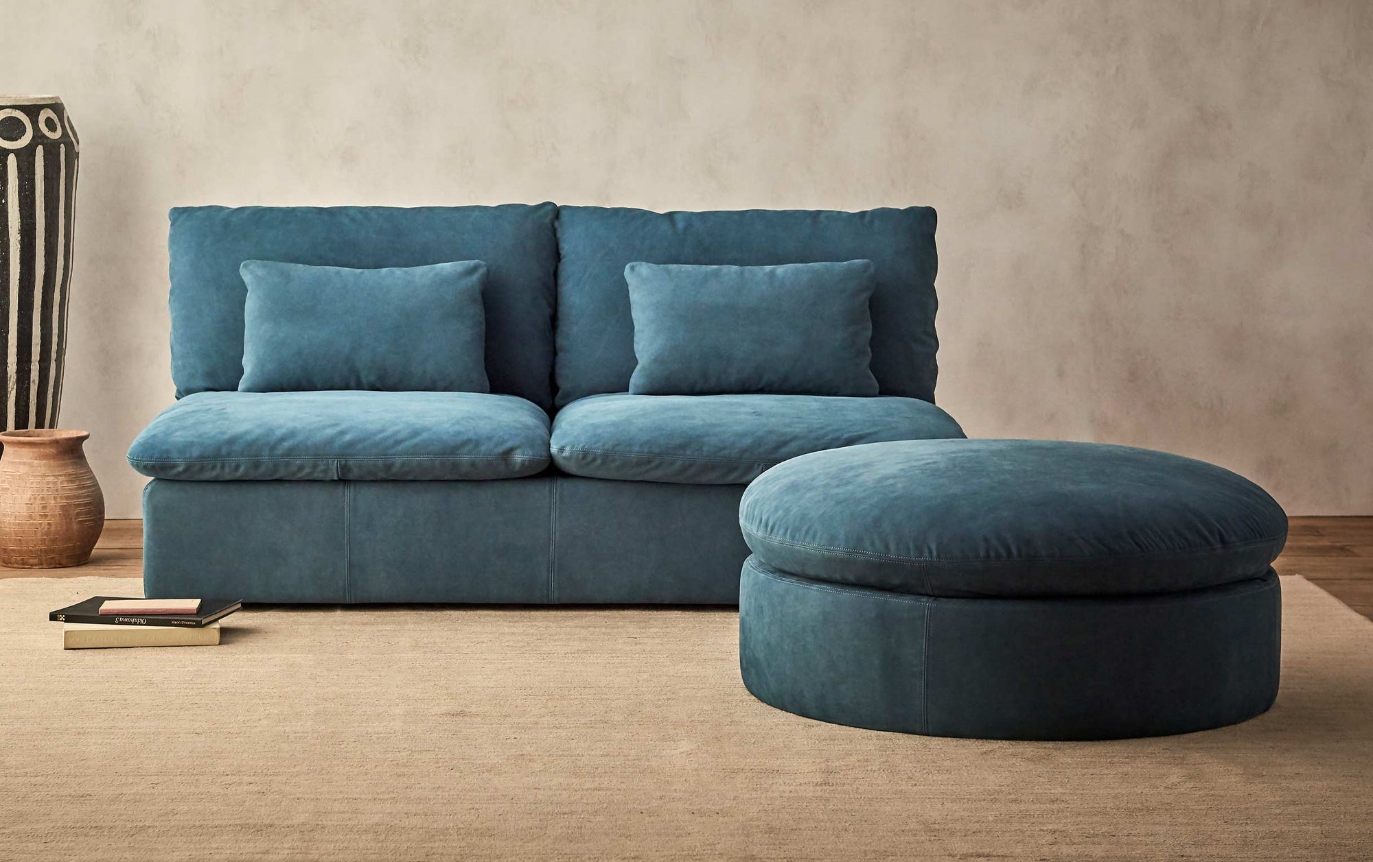 Aria Round Leather Ottoman in North Atlantic, a dark teal blue Meridian Leather placed on a beige rug in front of the Aria Leather Sofa in North Atlantic
