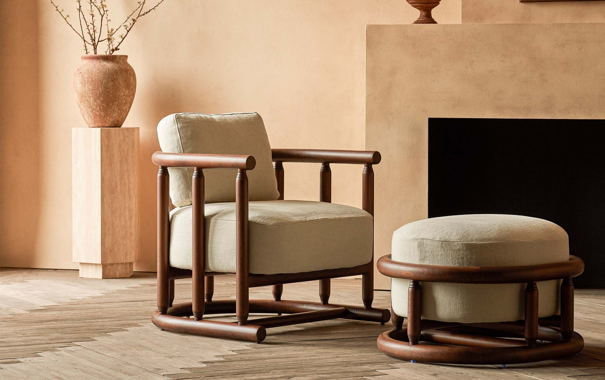Bene Chair in Dusky Ash, a deep, rich, molasses-colored 100% American ash, and Warm Oatmeal, a light warm beige Medium Weight Linen, placed in a warmly lit room with matching Bene Ottoman, in front of a fireplace and a potted plant on a stone pedestal