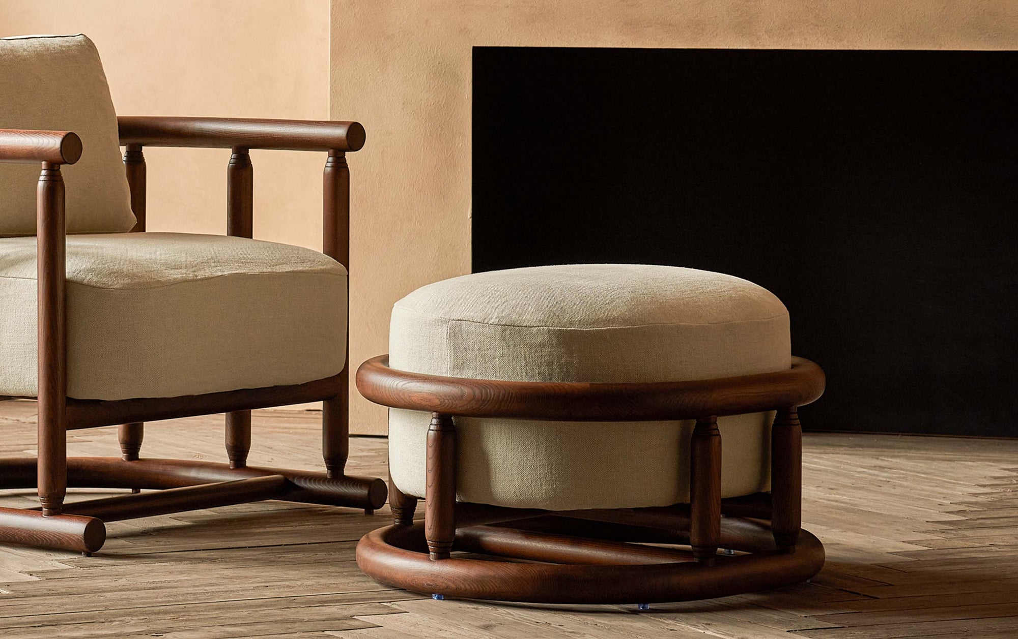 Bene Ottoman in Dusky Ash, a deep, rich, molasses-colored 100% American ash, and Warm Oatmeal, a light warm beige Medium Weight Linen, placed in a warmly lit room with matching Bene Chair in front of a fireplace