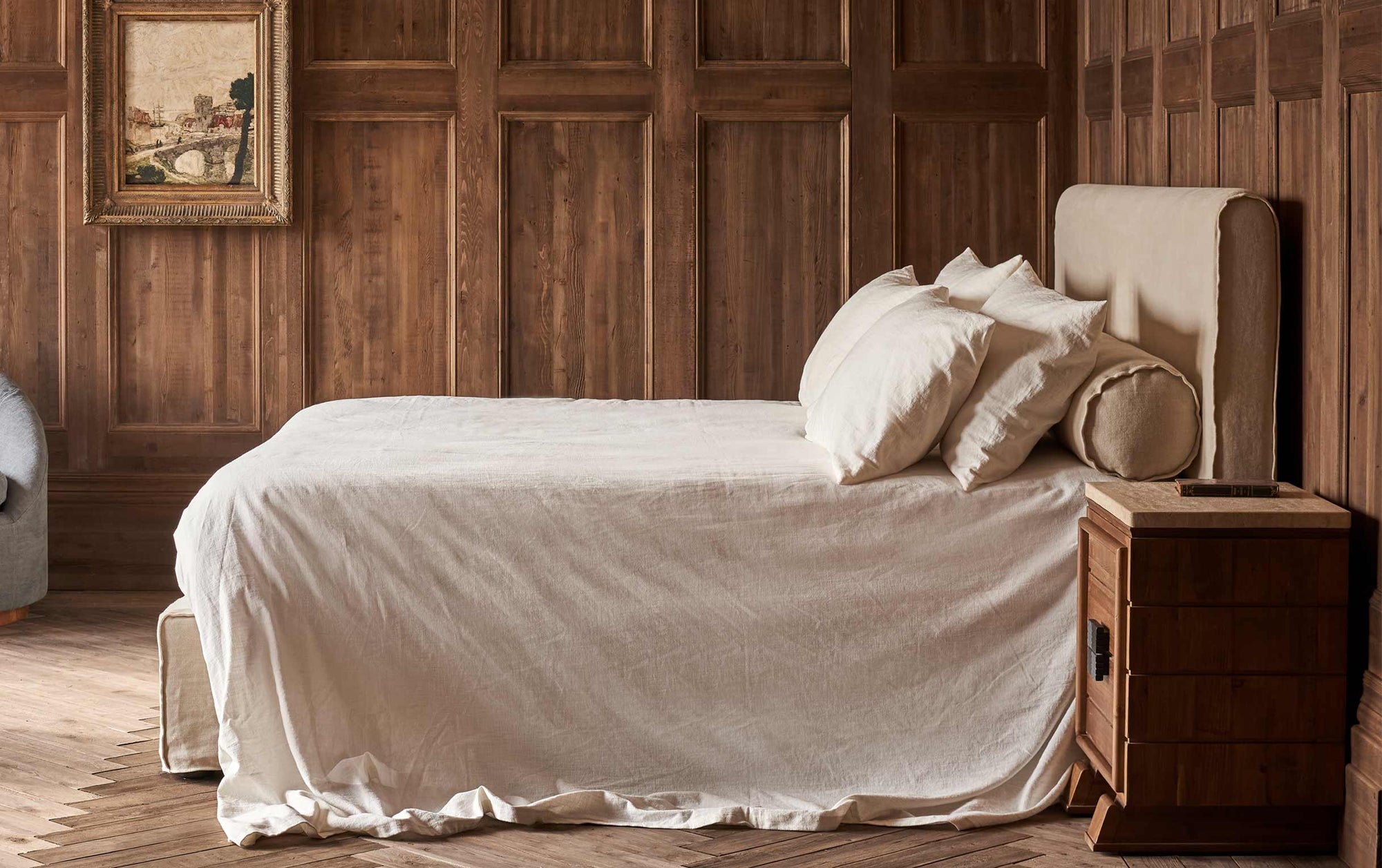 Profile of Camino King Bed in Warm Oatmeal, a light warm beige Medium Weight Linen, placed in a wood paneled room with a wooden nightstand