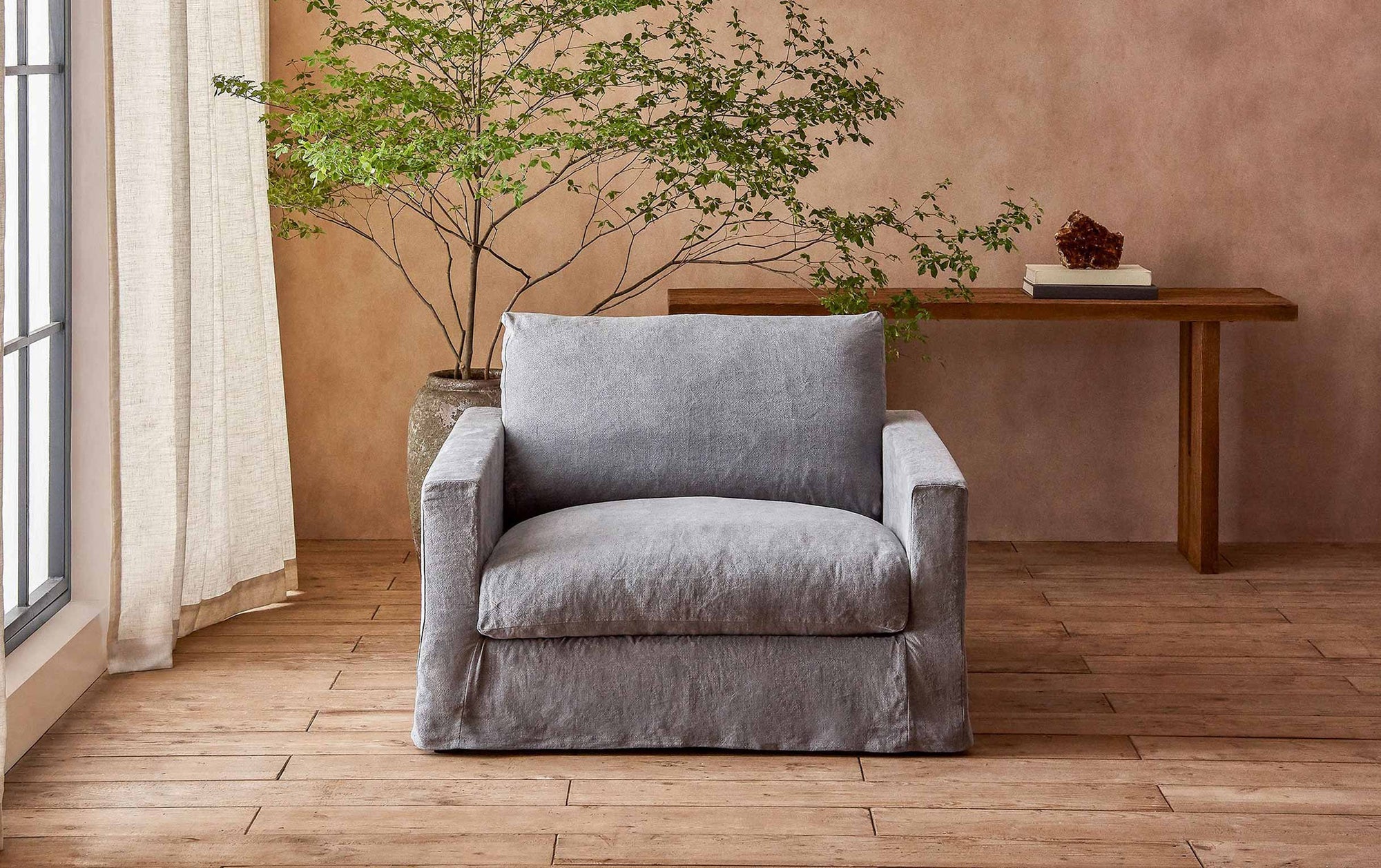 Devyn Chair in Ink Cap, a medium cool grey Light Weight Linen, placed in a warmly lit room in front of a Kai Console Table and a potted tree