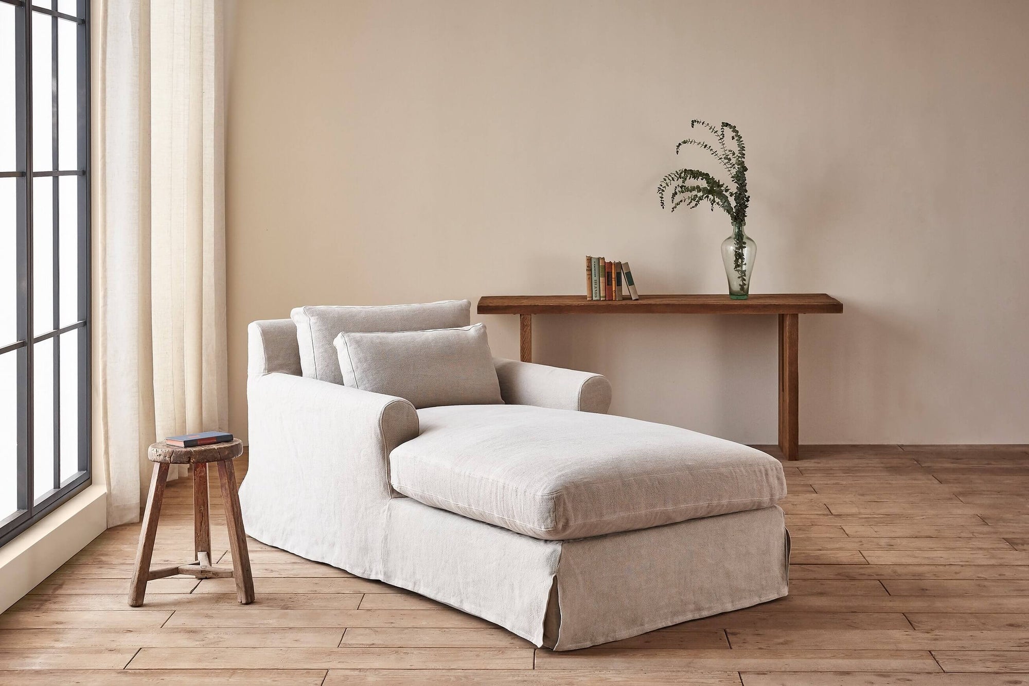Elias Daybed in Jasmine Rice, a light warm greige Medium Weight Linen, placed in a sunlit room with a wooden stool and console table decorated with books