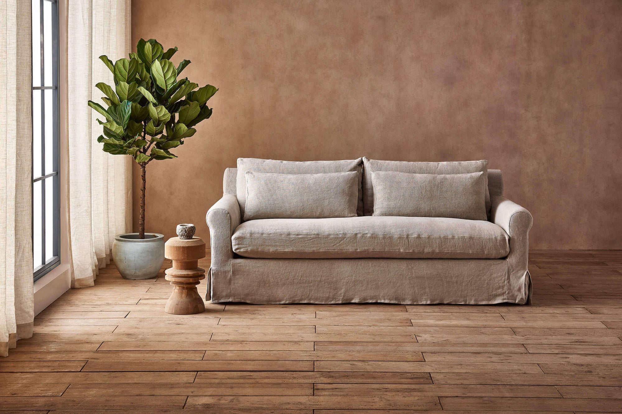 Elias Sofa in Jasmine Rice, a light warm greige Medium Weight Linen, next to a wooden end table with a large potted plant in the corner