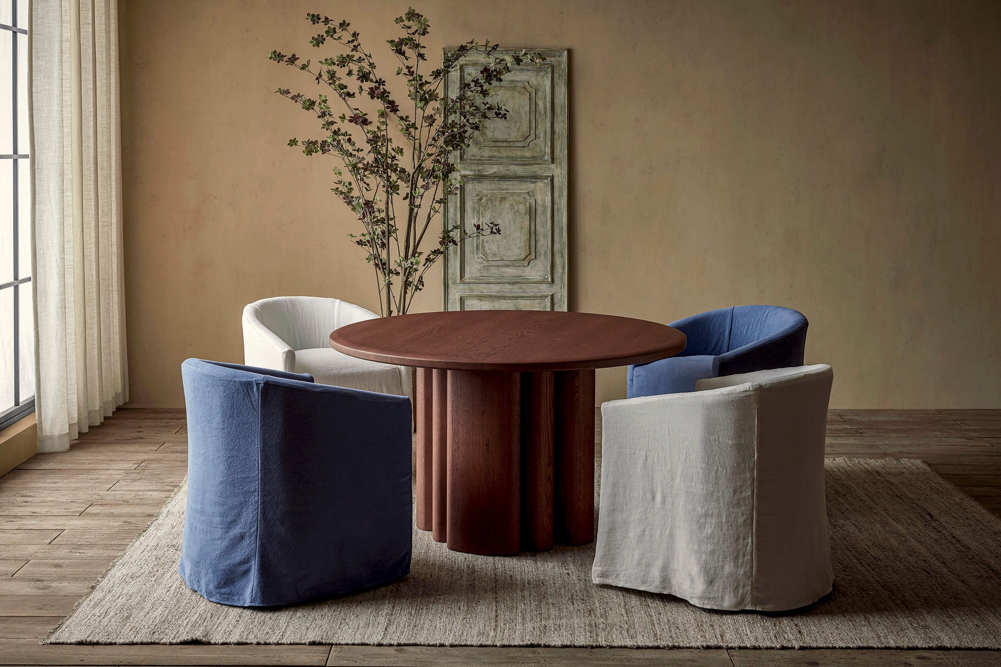 Four Ziki Dining Chairs in various colors placed on a rug around the Enzo Dining Table in Spiced Oak, in front of a decorative wooden art piece and a potted tree.