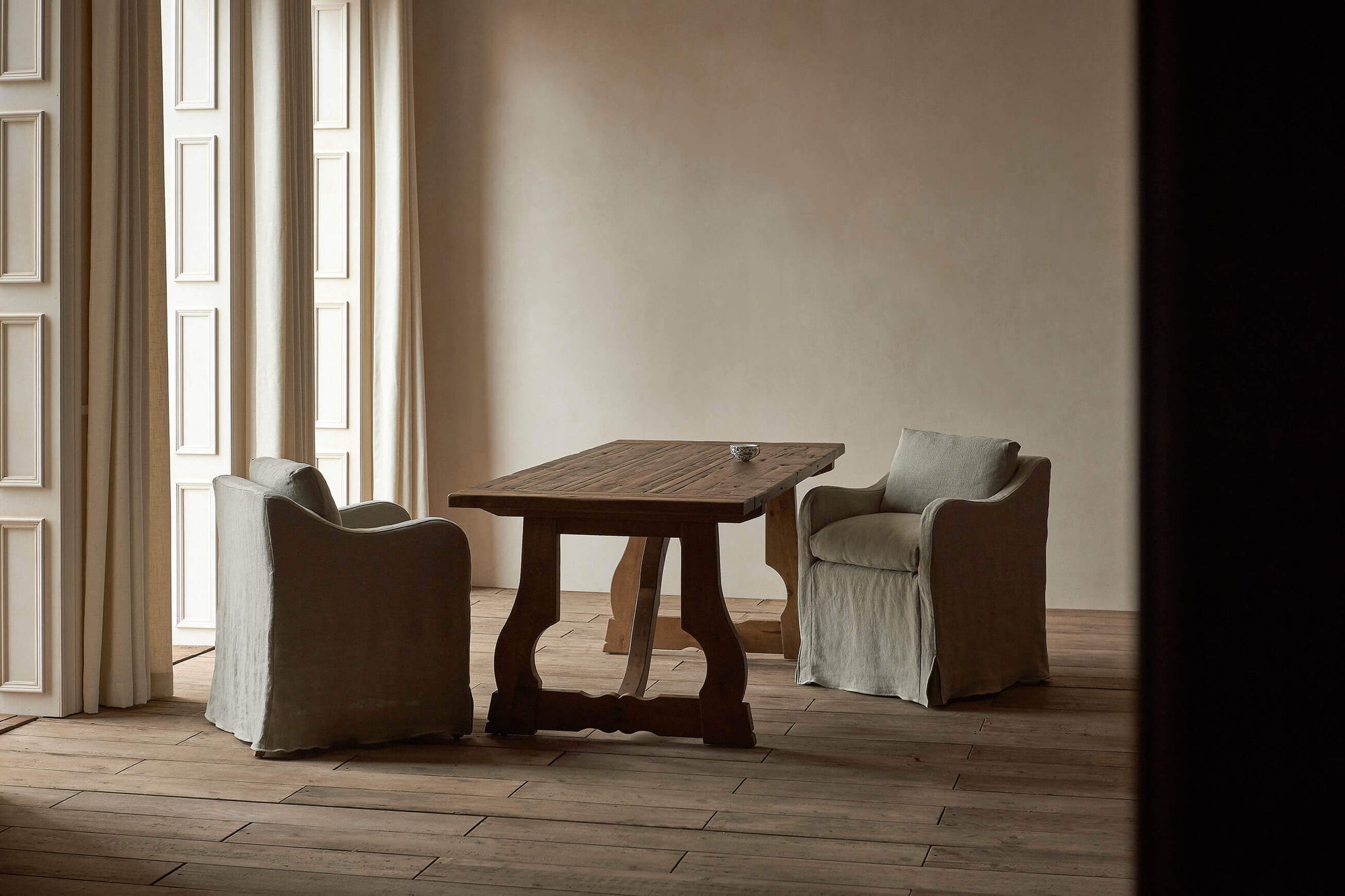 A Leona Dining Table in Heritage Pine is placed in a dining room with a set of two Esme Dining Chairs in Medium Weight Linen Jasmine Rice