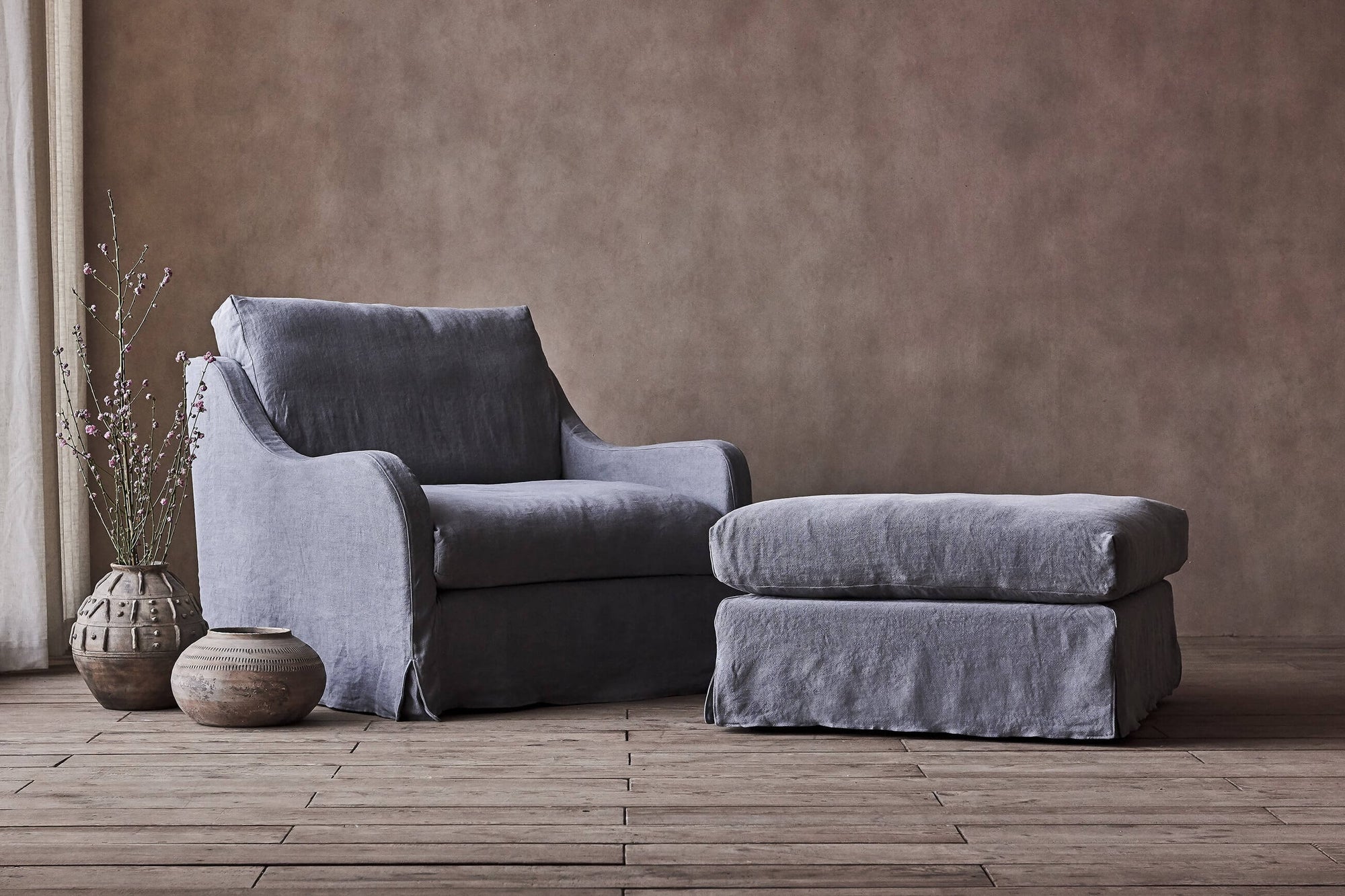 Esmé Chair Ottoman in Ink Cap, a medium cool grey Light Weight Linen, placed in a room alongside the Esmé Chair and potted plants on the floor