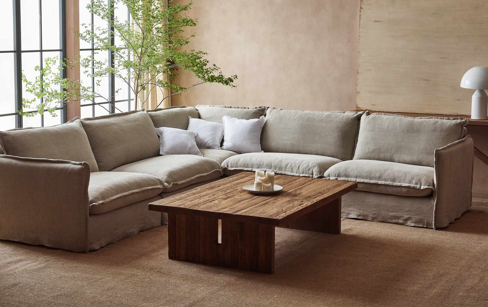 Kai Coffee Table in 100% Reclaimed Austrian Heritage Oak, a medium, bosc pear shade with earthy undertones, placed in front of the Neva Corner Sectional Sofa next to a large window and tall potted plant.