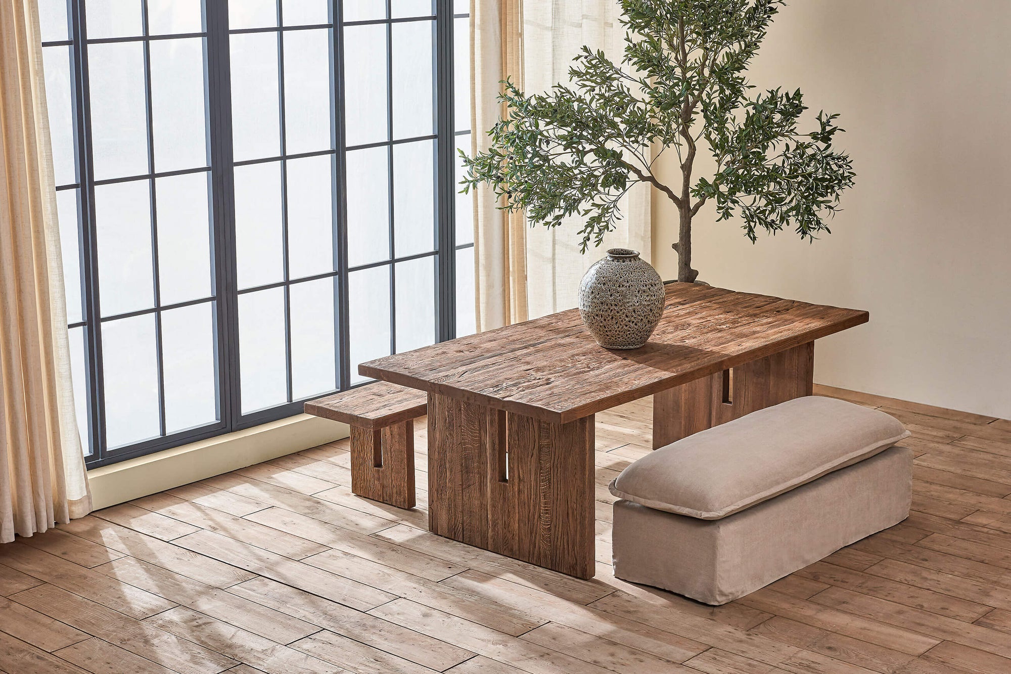 Kai Dining Table in 100% Reclaimed Austrian Heritage Oak, a medium, bosc pear shade with earthy undertones, placed in a sunlit room with the Kai Dining Bench and Neva Dining Bench, decorated with a large grey vase.