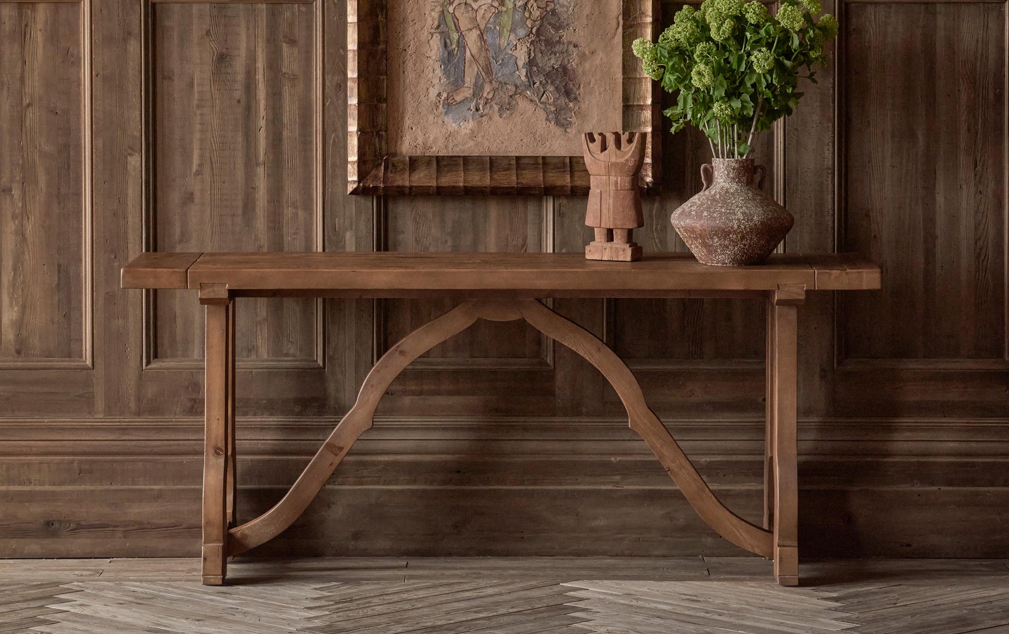 Leona Console Table in 100% Reclaimed Chinese Heritage Pine, a medium, bosc pear shade with earthy undertones, placed against a wood paneled wall, under a framed painting, and decorated with a wooden sculpture and a vase of flowers.
