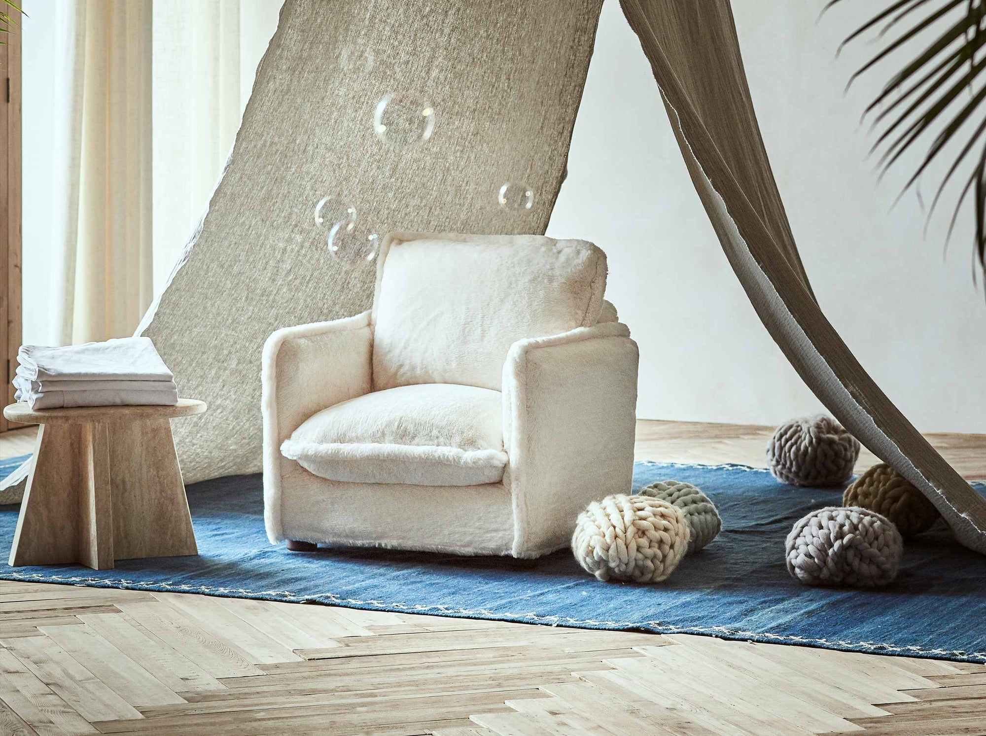 Little Neva Chair in Recycled Faux Fur Dandelion Poof placed on top of a blue rug between a side table and balls of yarn