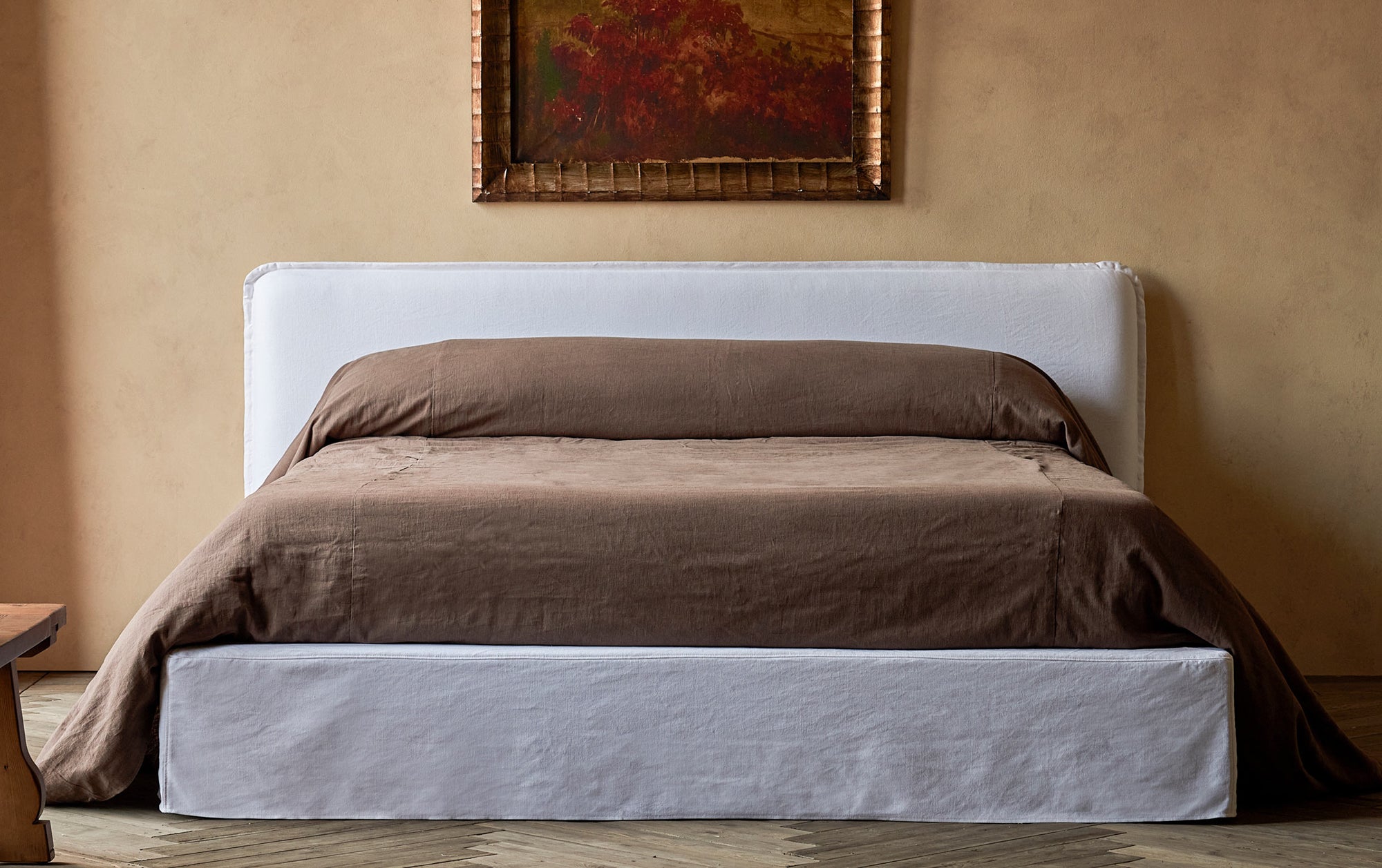 Neva California King Bed in Pacific Pearl, a cool, pure white Cotton Linen, place in a warmly lit room decorated with a vase of flowers on a Leona Bench and a painting on the wall.