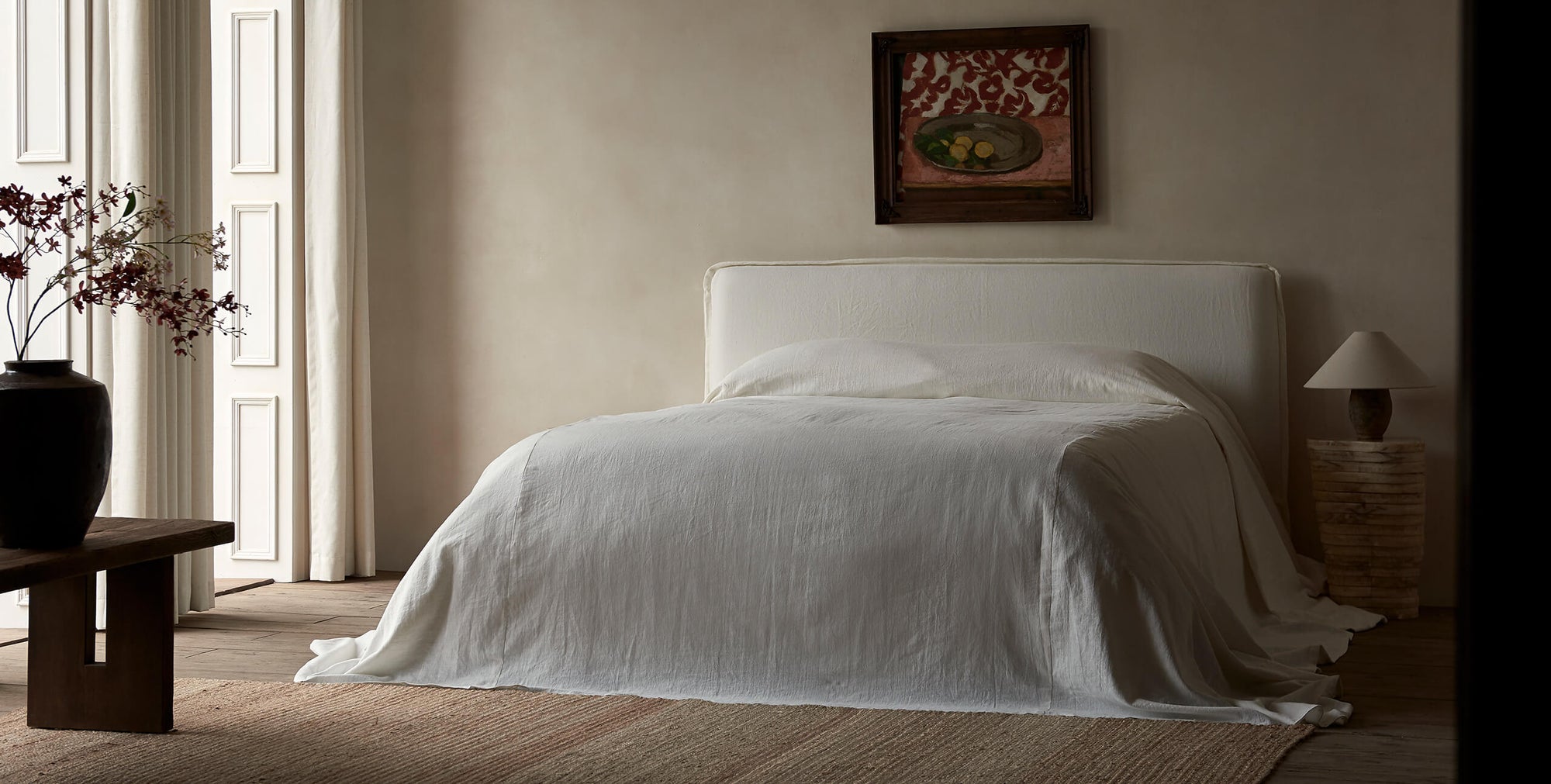 Neva Queen Bed in Water Lily, a white Light Weight Linen with warm undertones, placed in a decorated room with a stone side table and Kai Bench holding a plant in a large vase