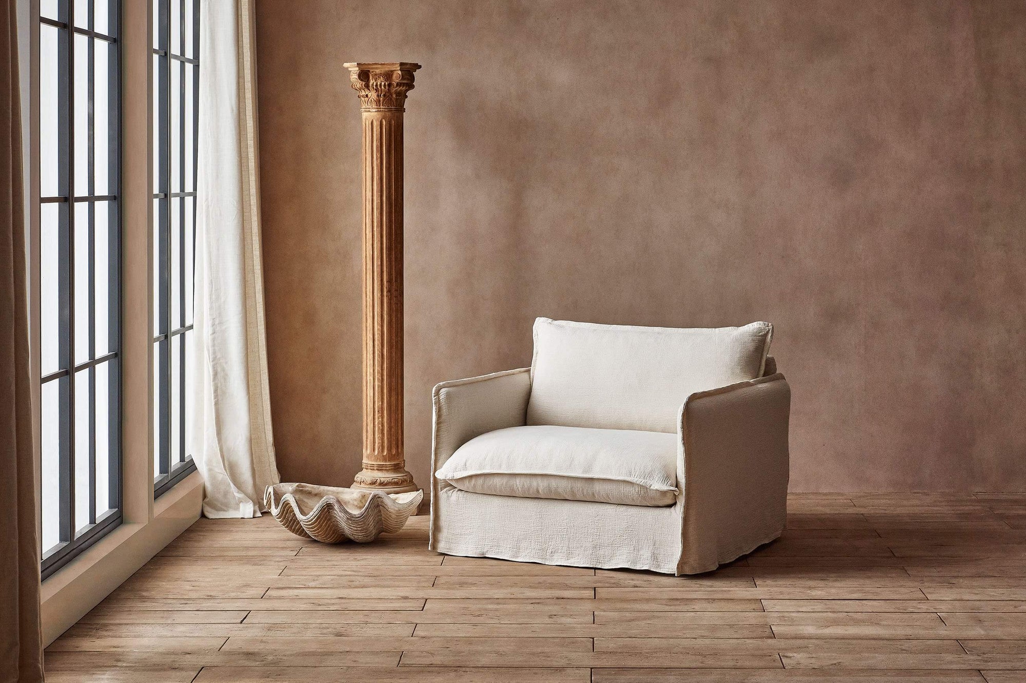 Neva Chair in Corn Silk, a light beige Washed Cotton Linen, placed in a brightly lit room with a column and a decprative shell sculpture.