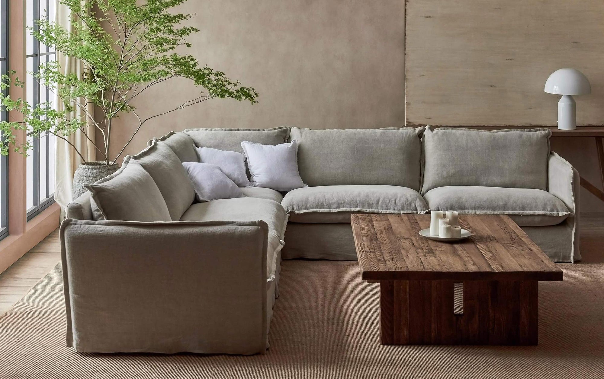 Neva Corner Sectional Sofa in Jasmine Rice, a light warm greige Medium Weight Linen, placed in a bright room with a Kai Coffee Table and a potted tree