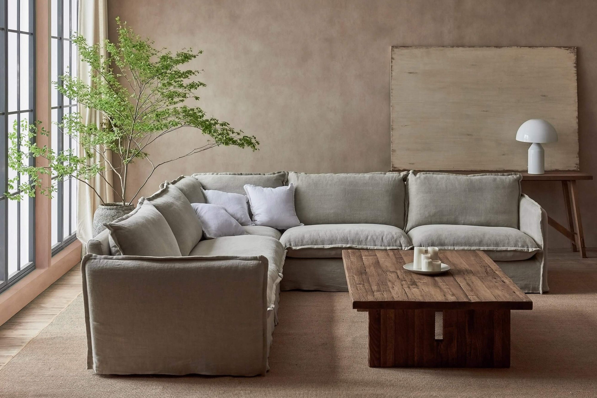 Neva Corner Sectional Sofa in Jasmine Rice, a light warm greige Medium Weight Linen, placed in a bright room with a Kai Coffee Table and a potted tree