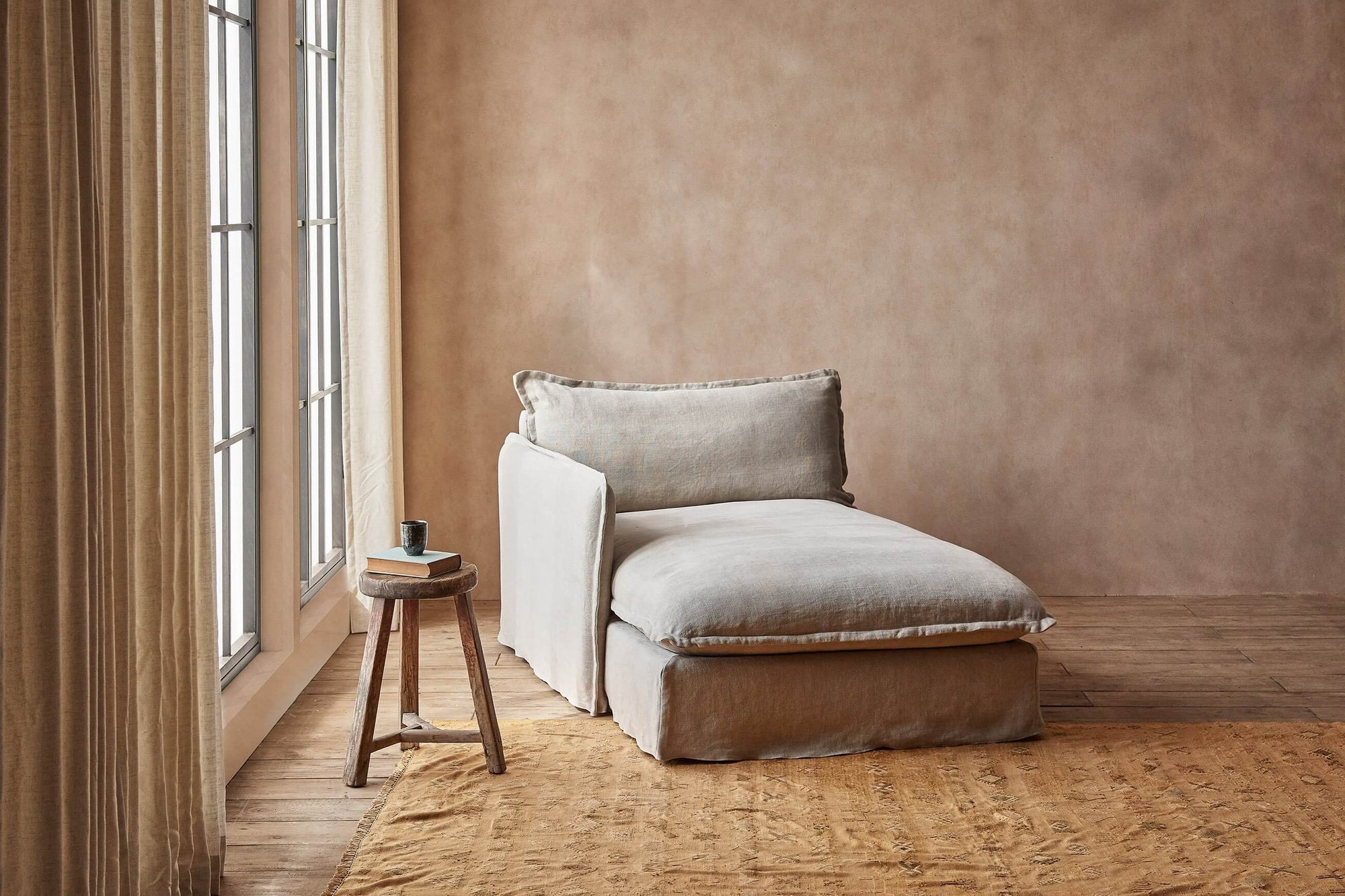 Neva Left Arm Facing Daybed in Jasmine Rice, a light warm greige Medium Weight Linen, placed in a room next to a large window and a stool holding a book and mug