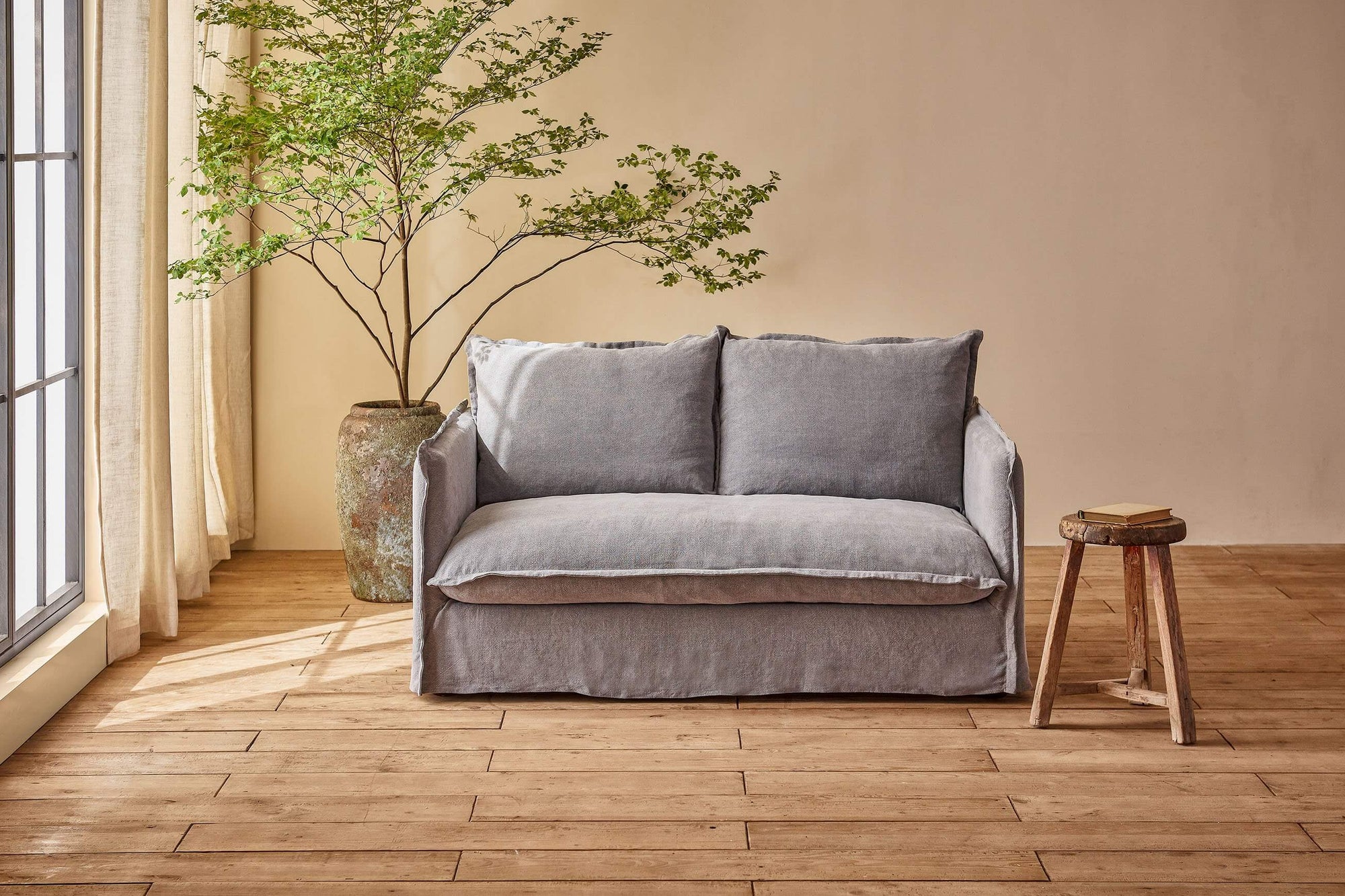 Neva 62" Loveseat in Ink Cap, a medium cool grey Light Weight Linen, placed in a sunlit room with a potted tree and a book on a stool