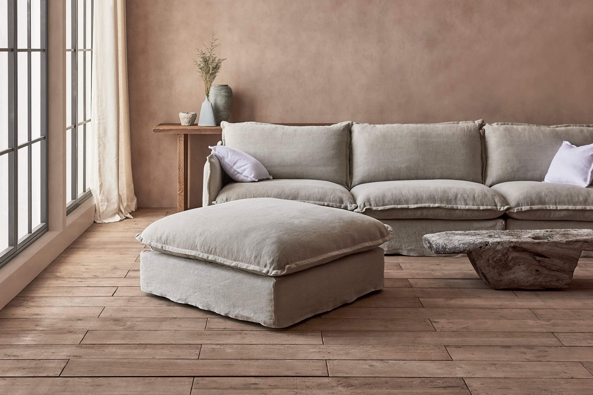 Neva Sectional Ottoman in Jasmine Rice, a light warm greige Medium Weight Linen, placed in a bright room in front of a Neva Sectional and a thin stone coffee table