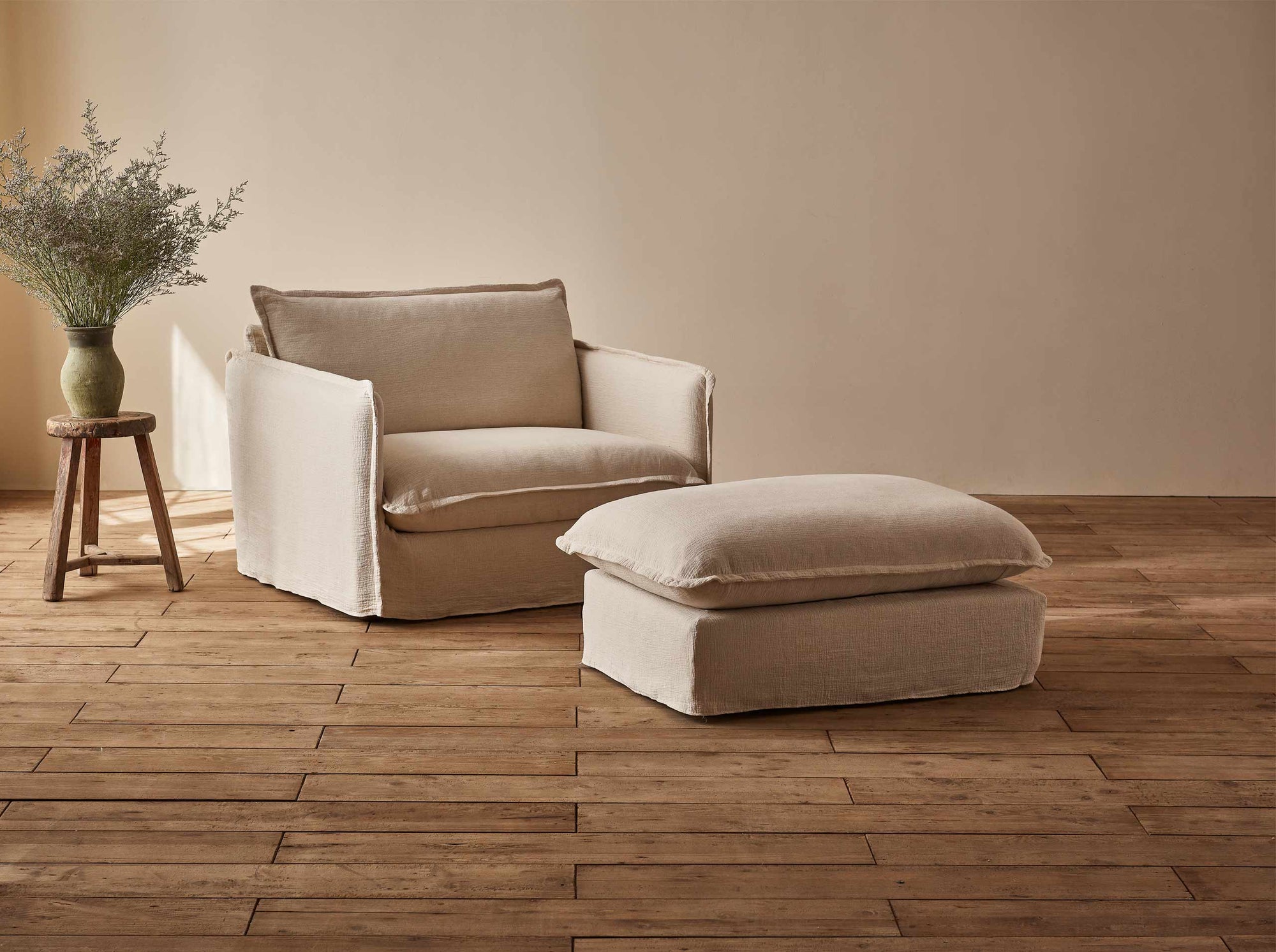 Neva Chair Ottoman in Corn Silk, a light beige Washed Cotton Linen, placed in front of a Neva Chair and a stool holding a vase of wildflowers