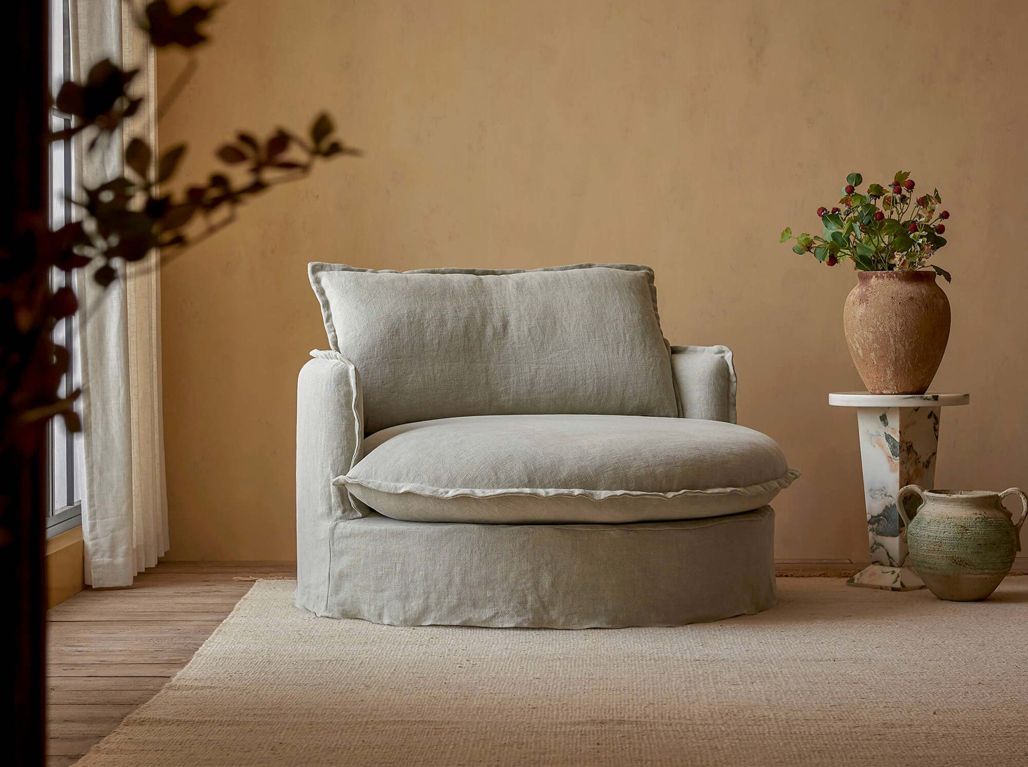 Neva Round Daybed in Jasmine Rice, a light warm greige Medium Weight Linen, placed in a sunlit room next to flowers in vases on a side table