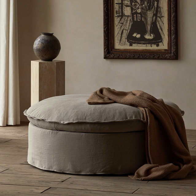 Neva Round Ottoman in Oat Flour, a medium taupe Light Weight Linen, with a brown throw blanket