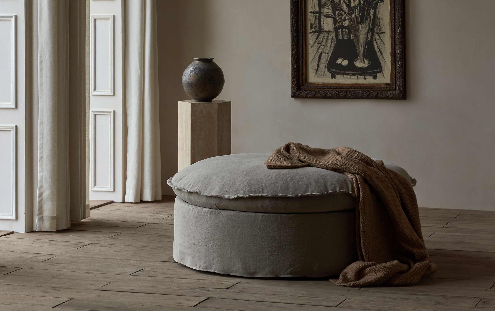 Neva Round Storage Ottoman in Oat Flour, a medium taupe Light Weight Linen, with a brown throw blanket, placed in a room decorated with art and a vase