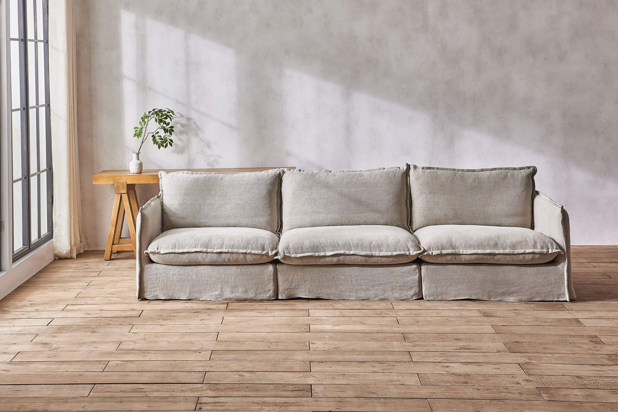 Neva Sectional Sofa in Jasmine Rice, a light warm greige Medium Weight Linen, placed in a sunlit room in front of a Rylance Console Table with a small plant