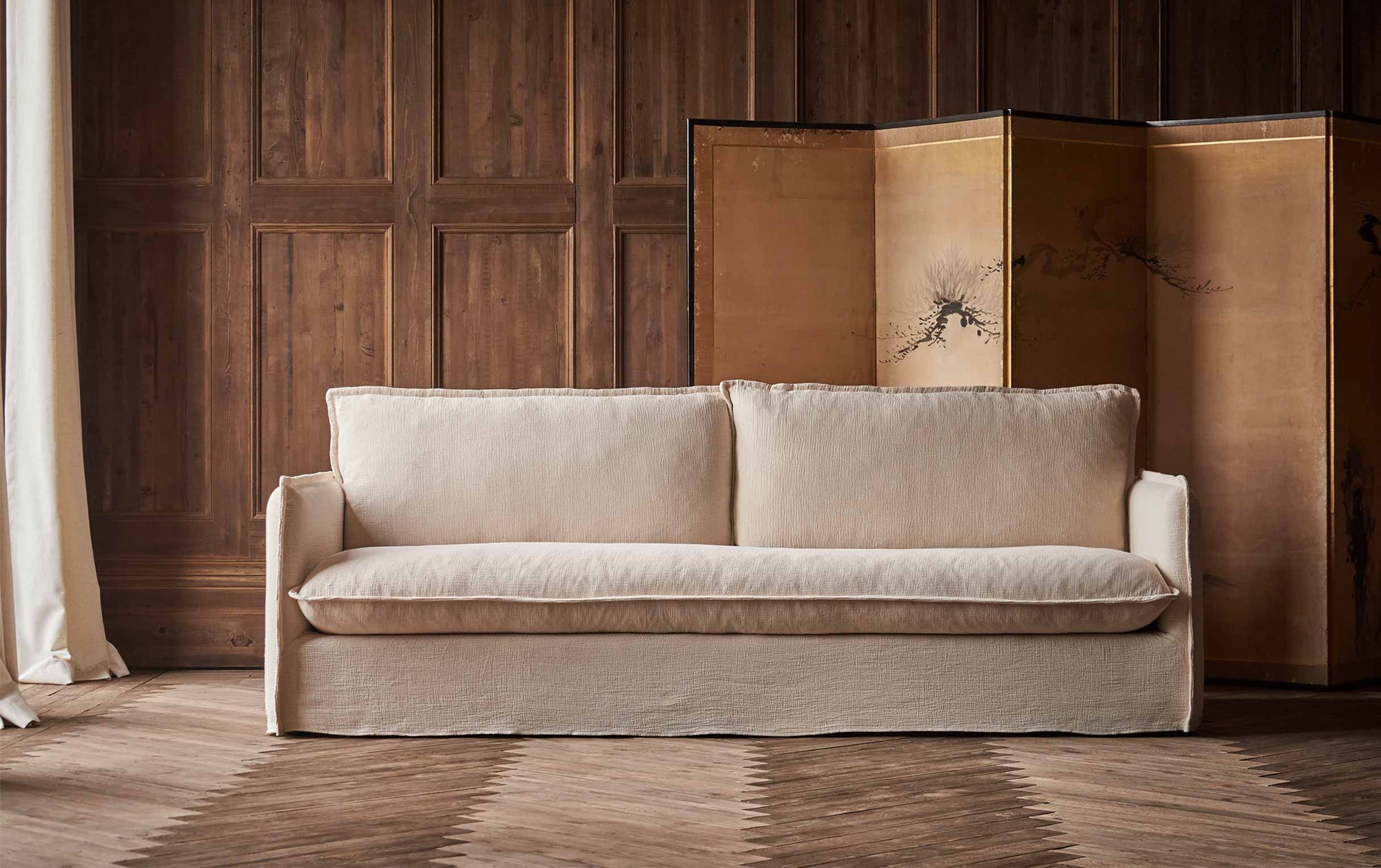 Neva 96" Sofa in Corn Silk, a light beige Washed Cotton Linen, placed in a wood-panelled room in front of a decorative folding screen