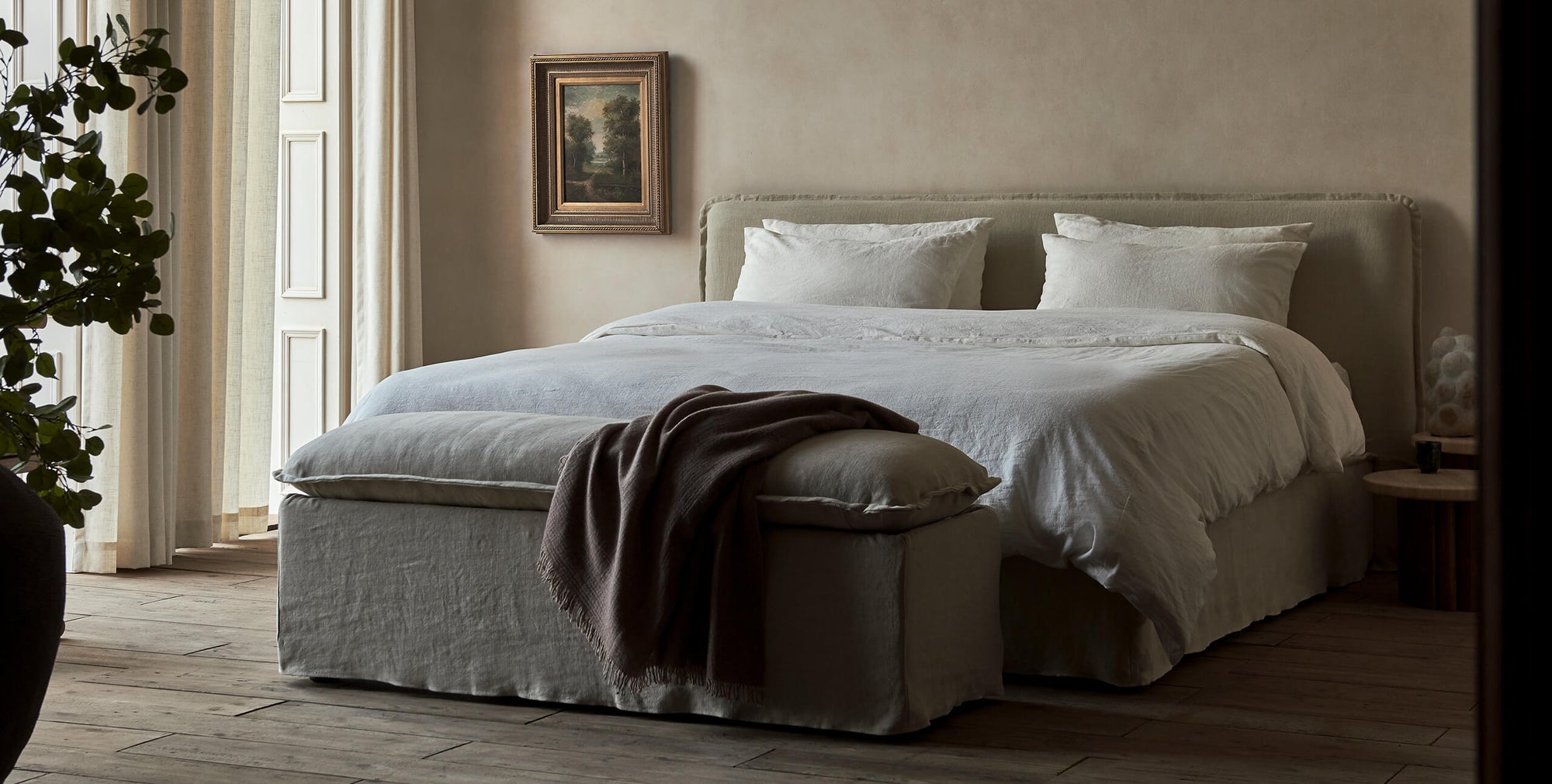 Neva Storage Bench in Jasmine Rice, a light warm greige Medium Weight Linen, with a brown throw blanket, placed at the end of a Neva King Bed in a decorated room