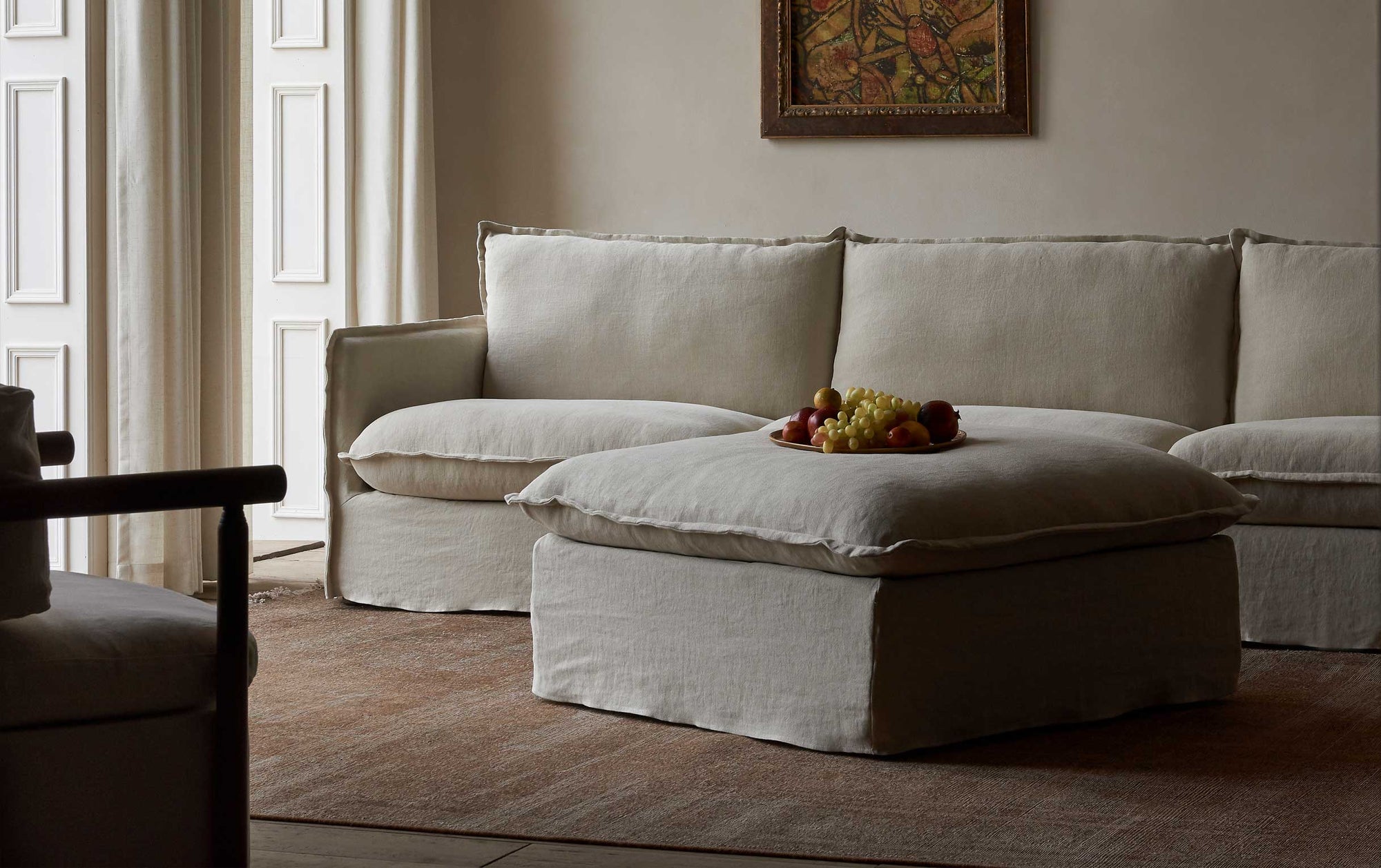 Neva Storage Ottoman in Jasmine Rice, a light warm greige Medium Weight Linen, placed in a decorated room in front of a Neva Sectional Sofa, with a tray of grapes on top
