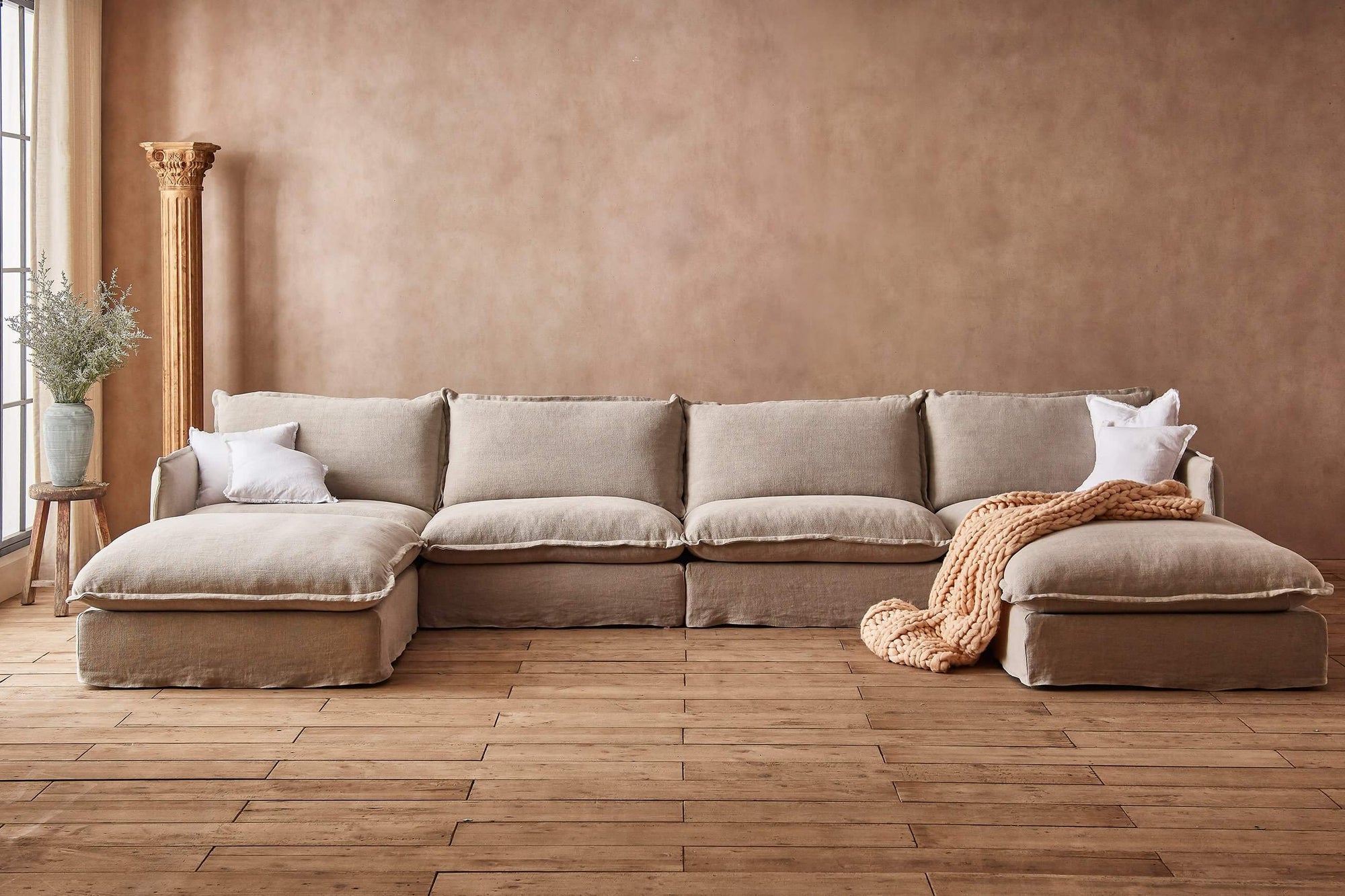 Neva U-Shape Sectional Sofa in Jasmine Rice, a light warm greige Medium Weight Linen, placed in a well-lit room with white throw pillows and a knit throw blanket, next to a plant on a stool.