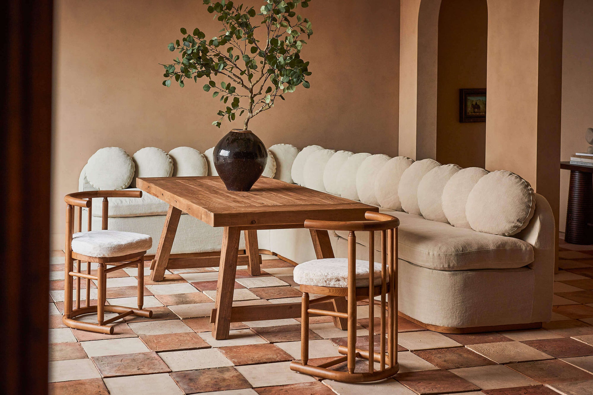 In a sunlit dining room, an Olea Banquette in Medium Weight Linen Warm Oatmeal surrounds the Fasso Dining Table in Heritage Pine and a set of 2 Bene Dining Chairs in Recycled Faux Fur Dandelion Poof