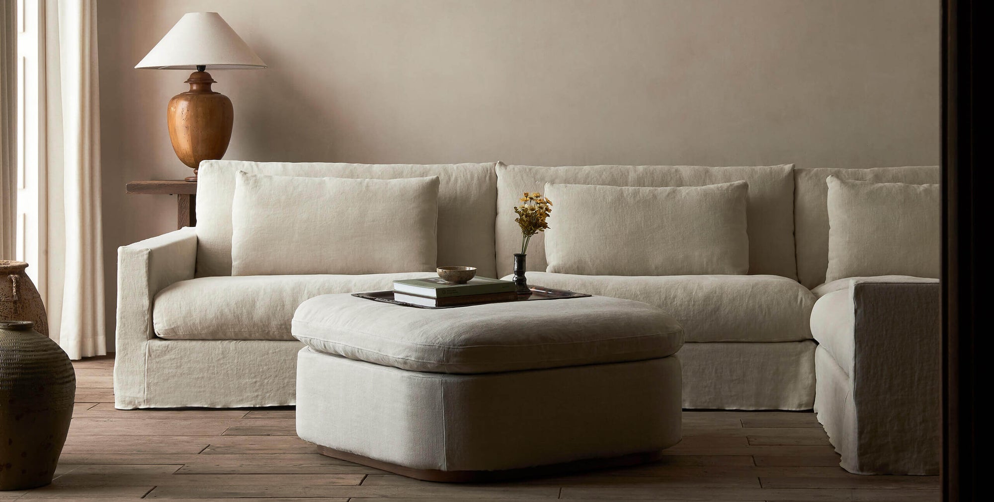 Olea Storage Ottoman in Warm Oatmeal, a light warm beige Medium Weight Linen, in front of a Devyn L-Shaped Sectional