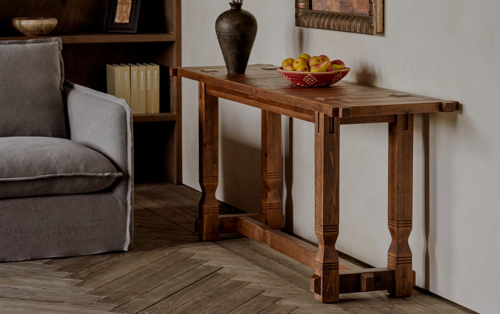 Ora Console Table in 100% Reclaimed Chinese Heritage Pine, a medium, bosc pear shade with earthy undertones, placed in a room with a Neva Sofa, in front of a bookcase, and decorated with a vase and a bowl of pears.
