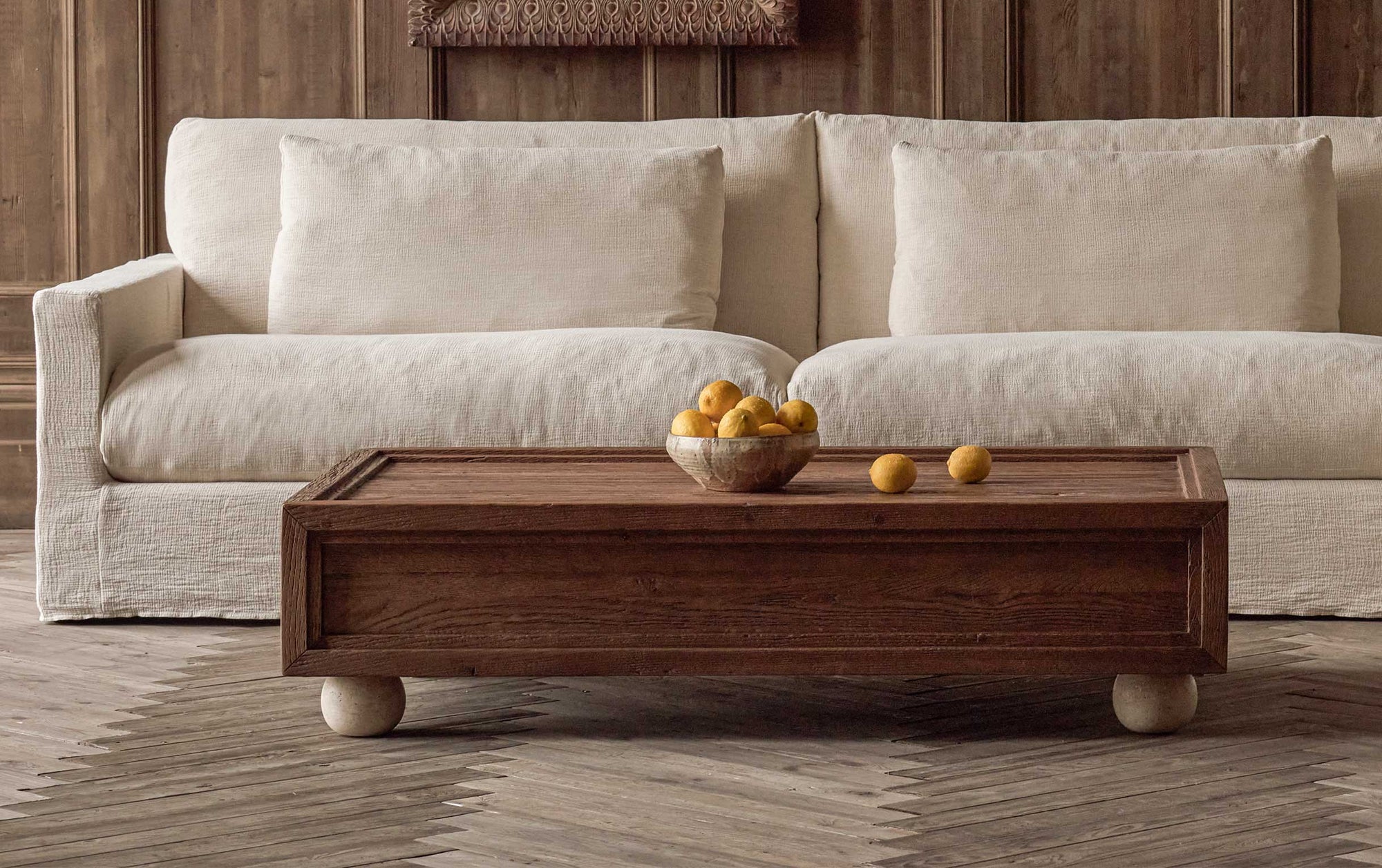 Rho Coffee Table in 100% Reclaimed Austrian Heritage Oak, a medium, bosc pear shade with earthy undertones, placed in a wood paneled room in front of the Devyn Sofa, topped with a bowl of citrus fruit.