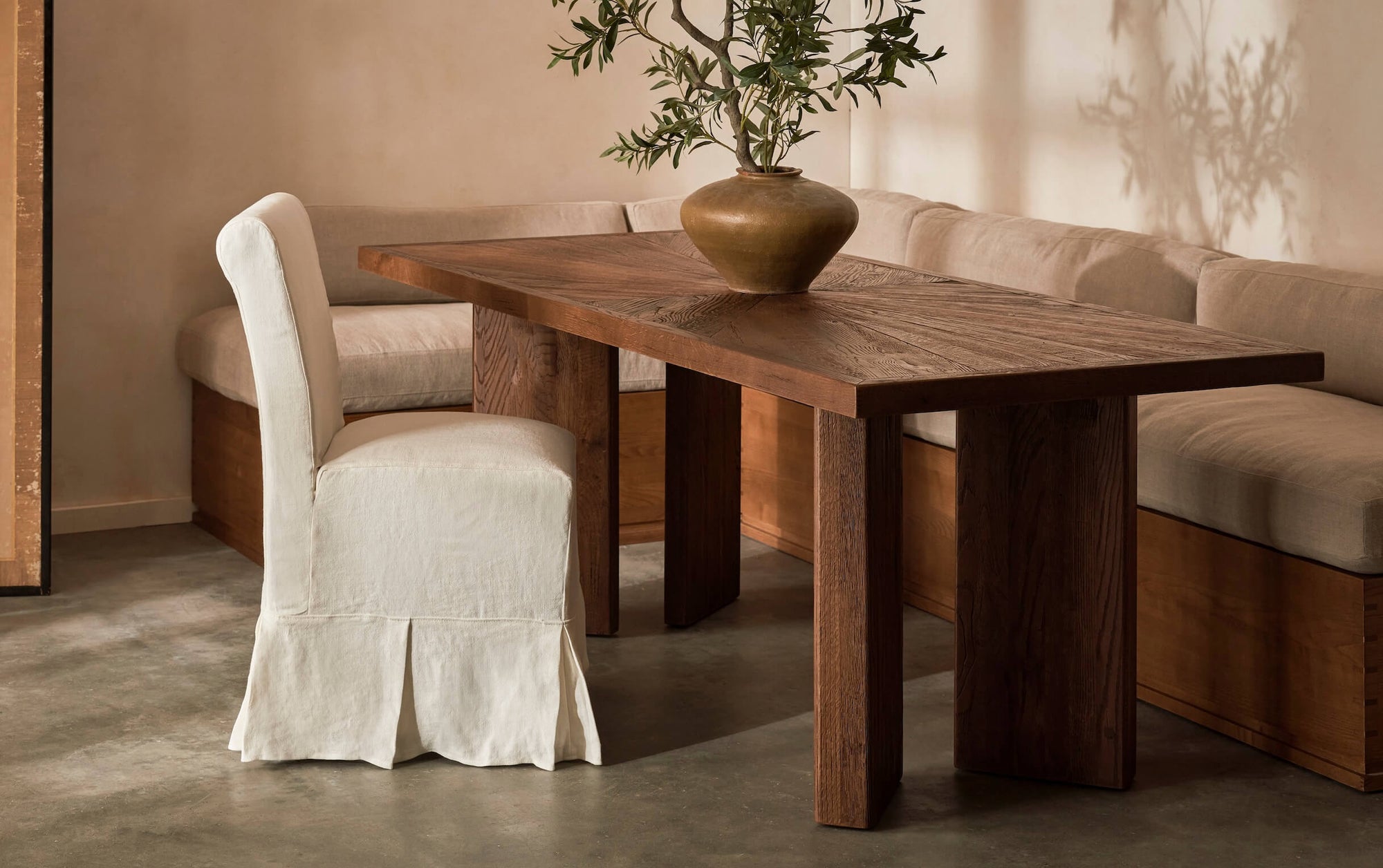Tango Dining Table in 100% Reclaimed Austrian Heritage Oak, a medium, bosc pear shade with earthy undertones, places in a sunlit room with a Monti Banquette and a Dalia Armless Dining Chair