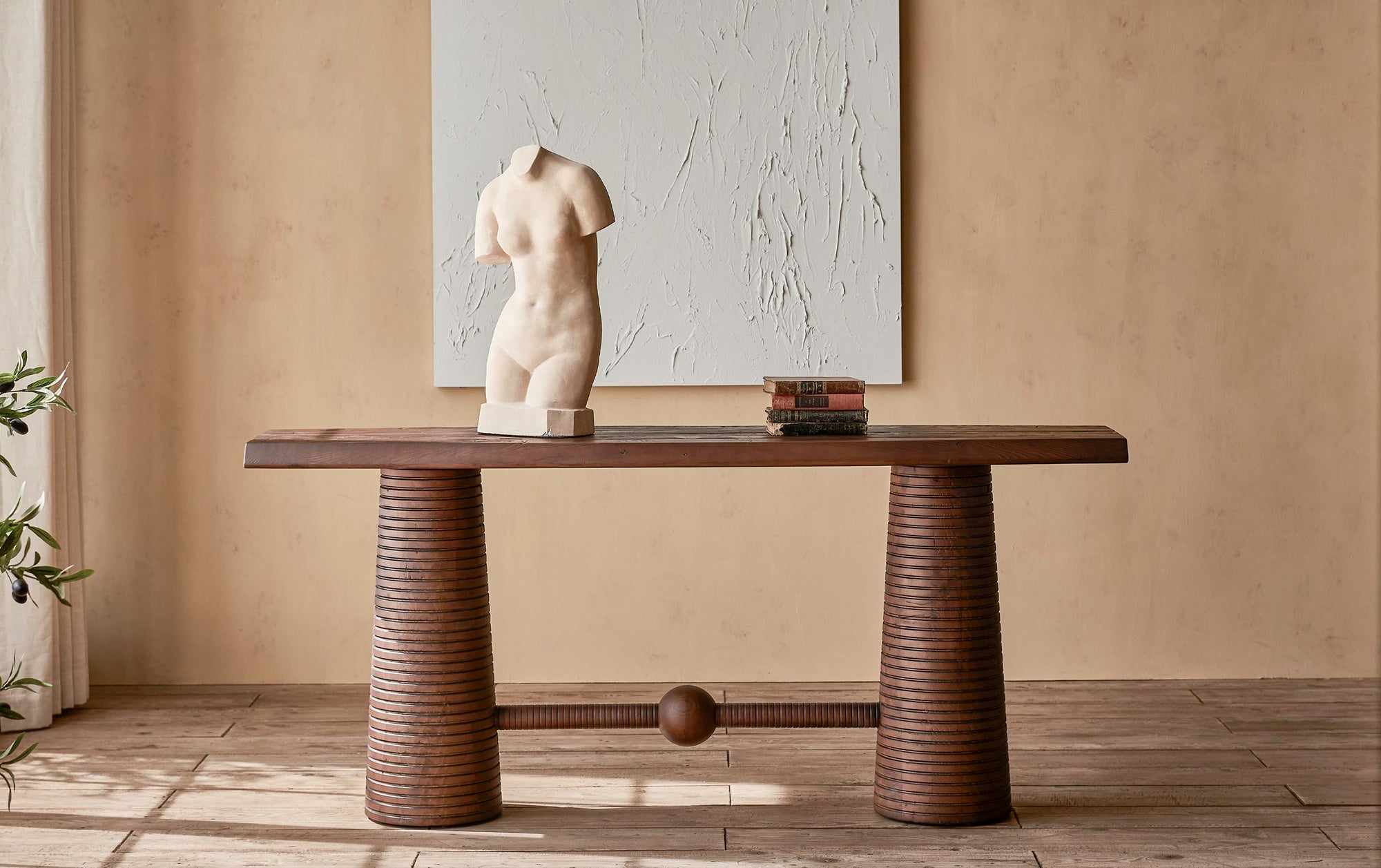 A feminine statue and stack of books on top of the Zenia Console Table in Dusky Pine, a deep, rich, molasses colored 100% Chinese pine, placed in a sunlit room in front of a large white painting.