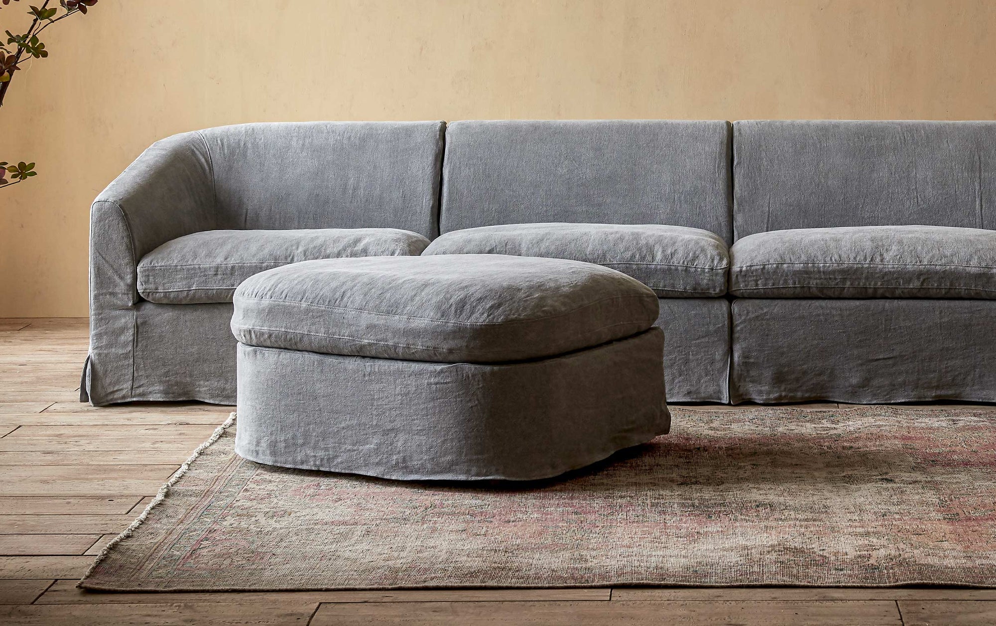 Ziki Sectional Ottoman in Ink Cap, a medium cool grey Light Weight Linen, placed on a rug in front of the Ziki Sectional Sofa