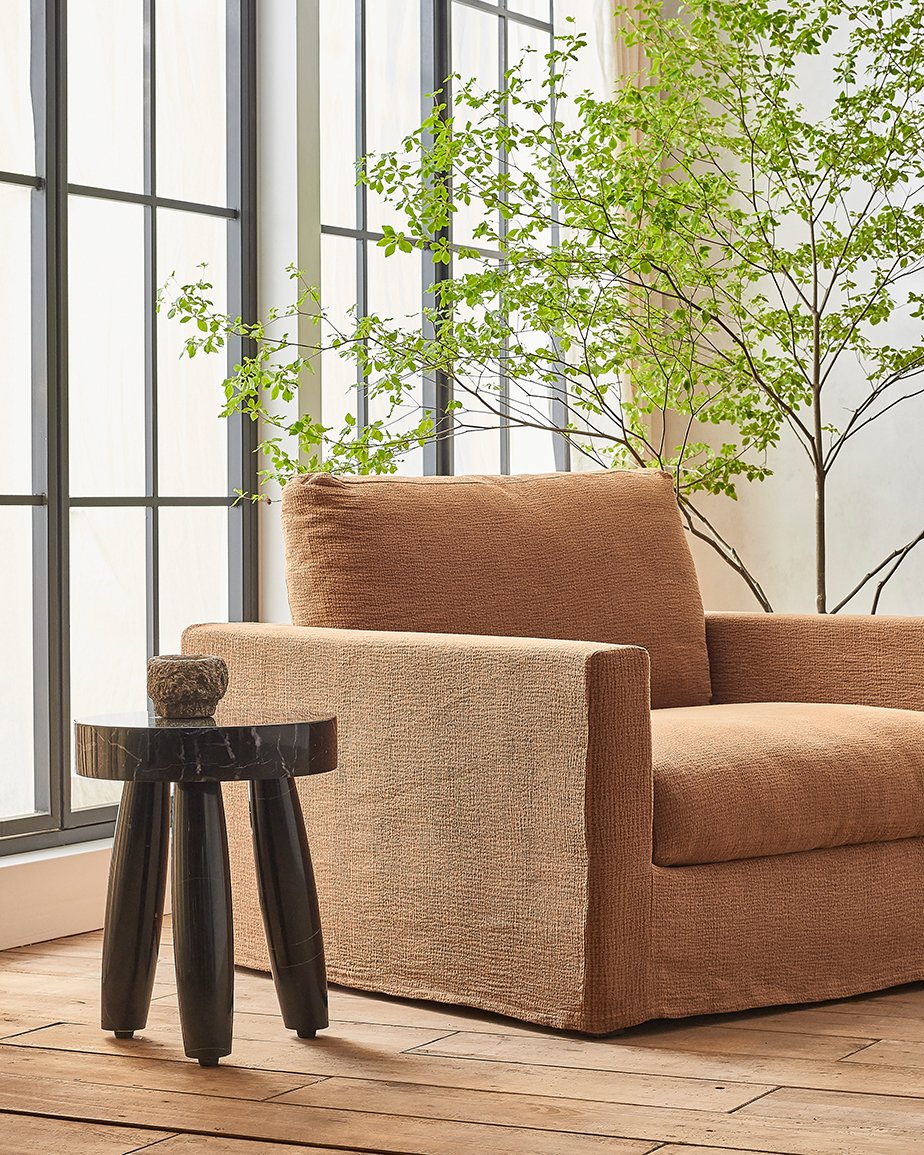 Devyn Chair in Sweet Potato, a copper brown Washed Cotton Linen, placed in a sunlit room between a stool and potted tree