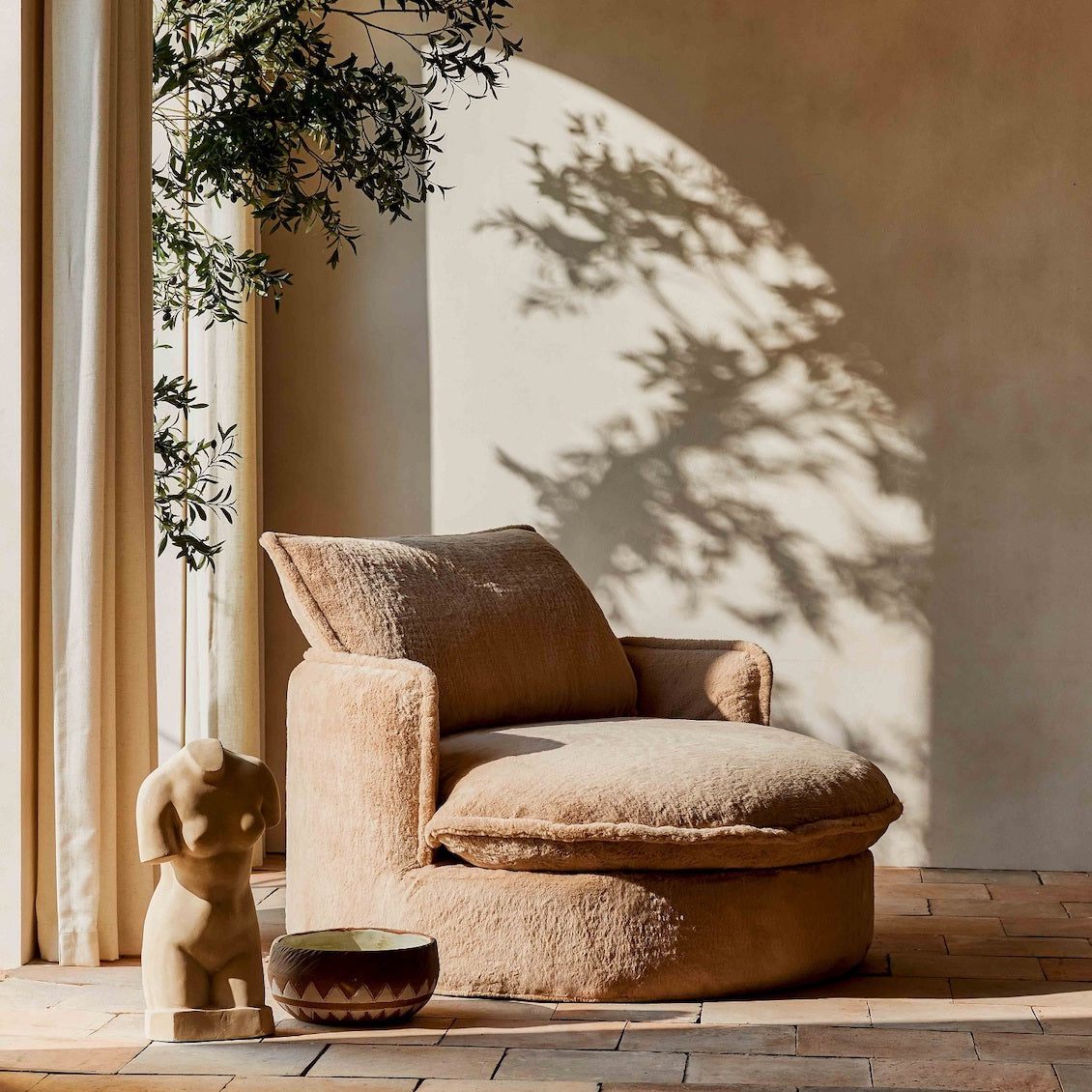 Neva Round Daybed in Pampas Flow, a light tan brown Recycled Faux Fur, placed in a sunlit room with a floor plant