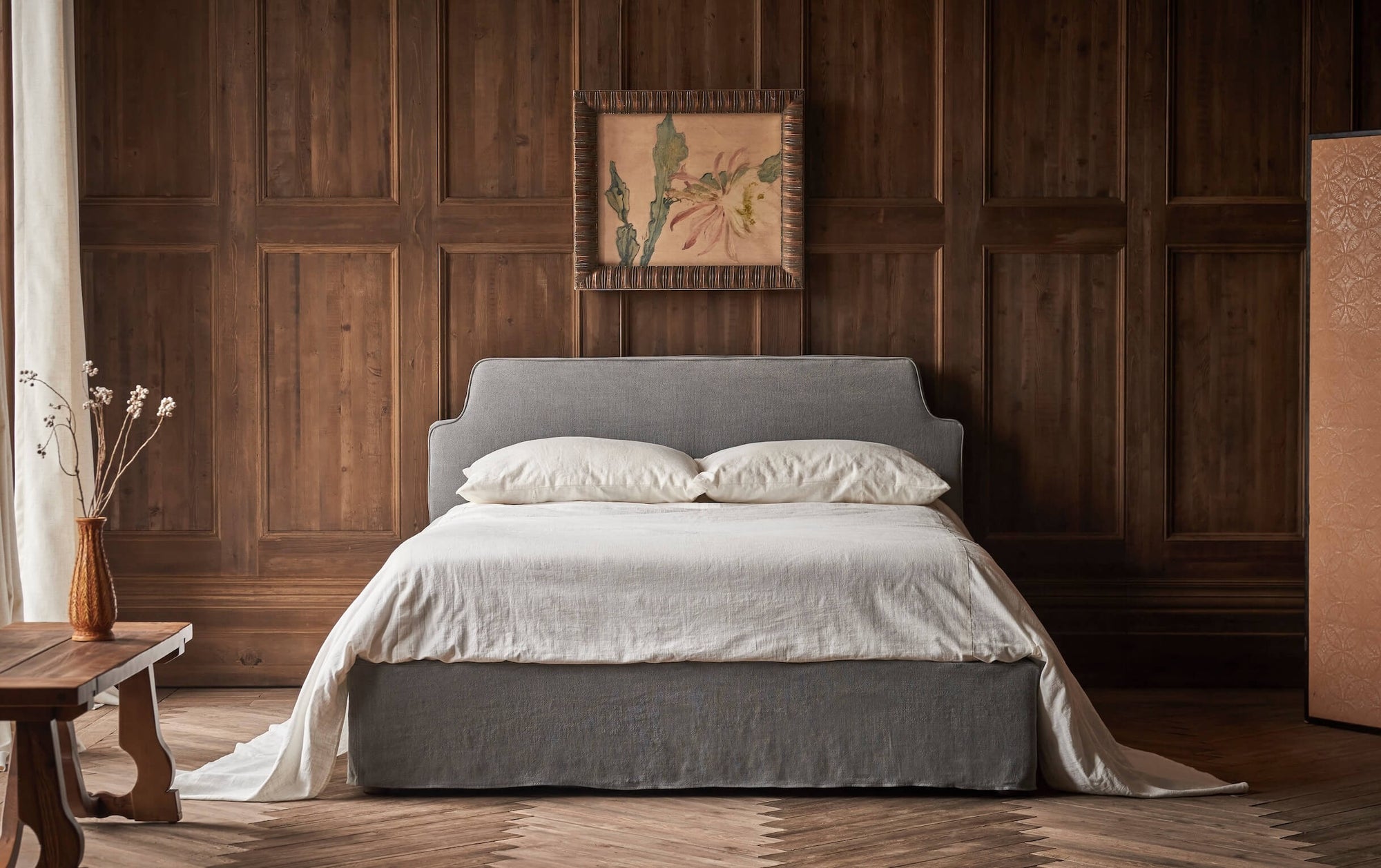 Amelia Bed in Ink Cap, a medium cool grey Light Weight Linen, made with white bed linens and placed In a wood paneled room with a Leona Dining Bench holding a vase of flowers.