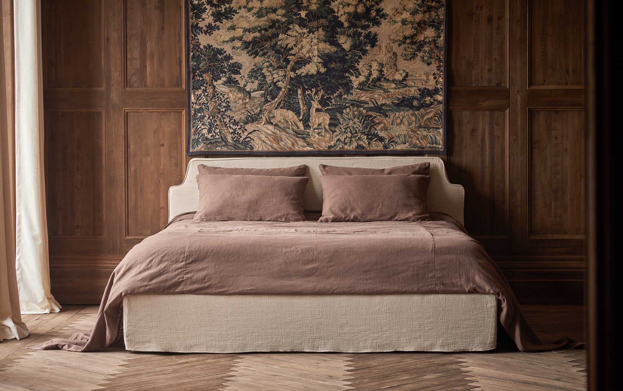 Amelia King Bed in Corn Silk, a light beige Washed Cotton Linen, made up in dusty mauve bedding, placed in a wood-paneled room with a large framed tapestry