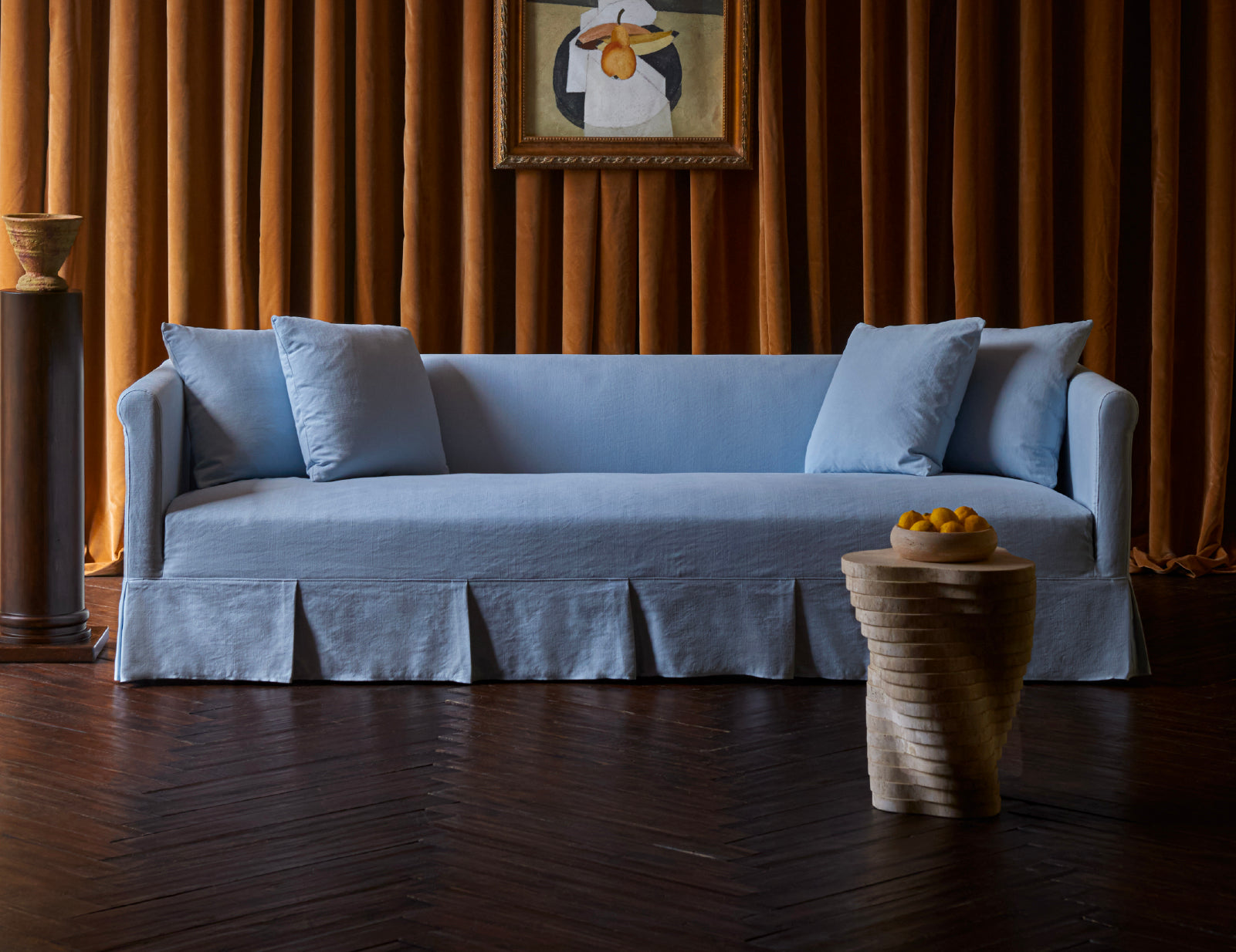 A Sixpenny Dalia sofa in salt water with a skirted base and matching back cushions, styled in a room with dark wood flooring and warm brown curtains. A Sixpenny Galata Travertine side table with a bowl of oranges sits in the foreground.