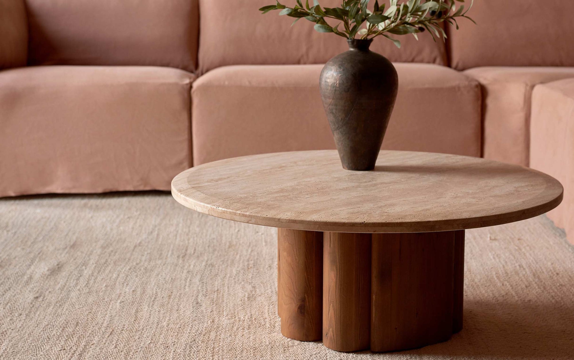 Enzo Coffee Table in Turkish Galata Travertine in a warm beige with gold and pink undertones. Various veining, color variations, and pitting throughout. Table base made from 100% Reclaimed Chinese Pine. Table is topped with a plant and placed in front of a Loula L-Shape Sectional in Nectarine Dream, on a beige rug.