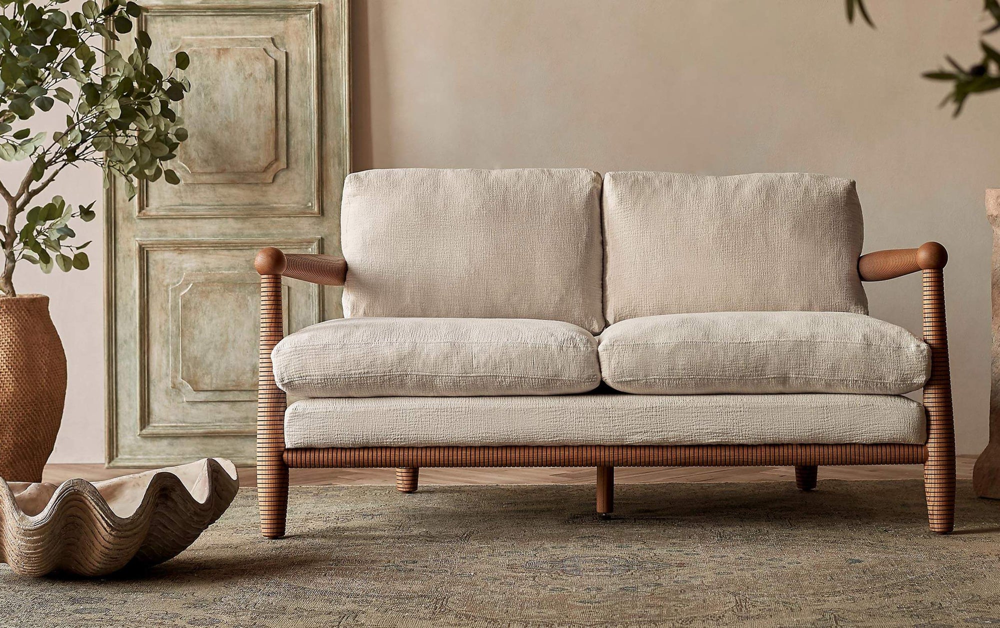 Gio Loveseat in a Dusky Ash wood frame with cushions in Corn Silk, a light beige Washed Cotton Linen, next to a clam sculpture and a large potted plant